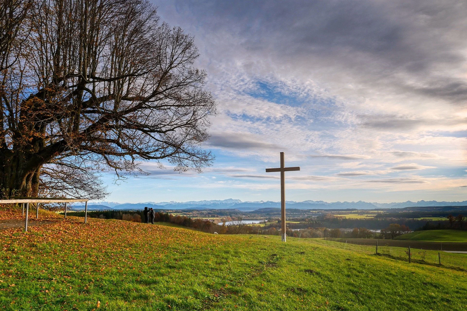 A scenic landscape features a large leafless tree on the left, a wooden cross on the right, and a couple walking hand-in-hand in the distance, with mountains and a cloudy sky in the background.