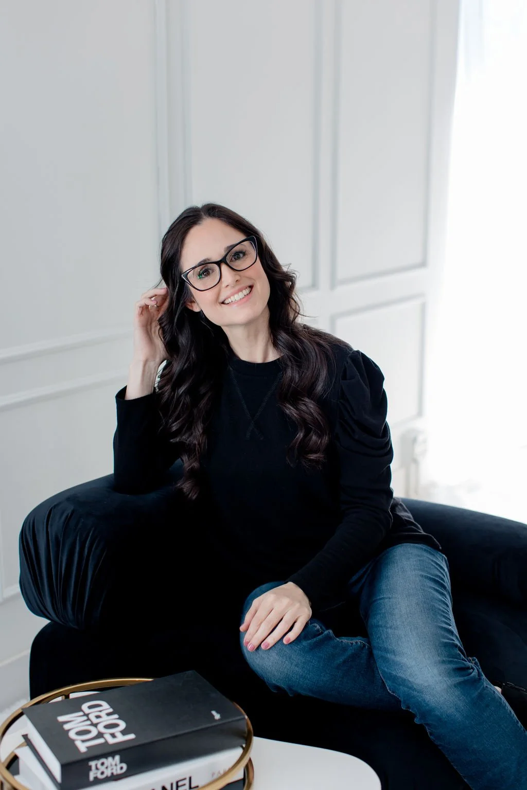A woman with long dark hair, glasses, and a big smile, sitting on a black sofa in a white-walled room, wearing a black top and jeans, with a book titled "Tom Ford" on a round table in front of her.
