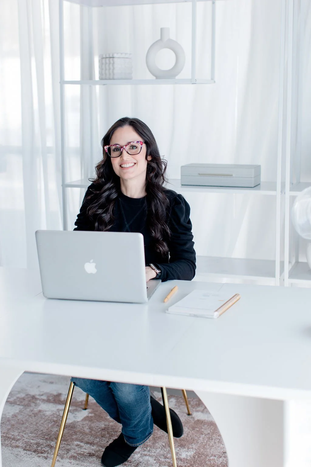 Melie is sitting at a white desk with a silver MacBook. She is smiling and wearing a black top. On the desk are a notebook, a pen, and some papers. Behind her is a white shelf with decorative objects and sheer curtains.