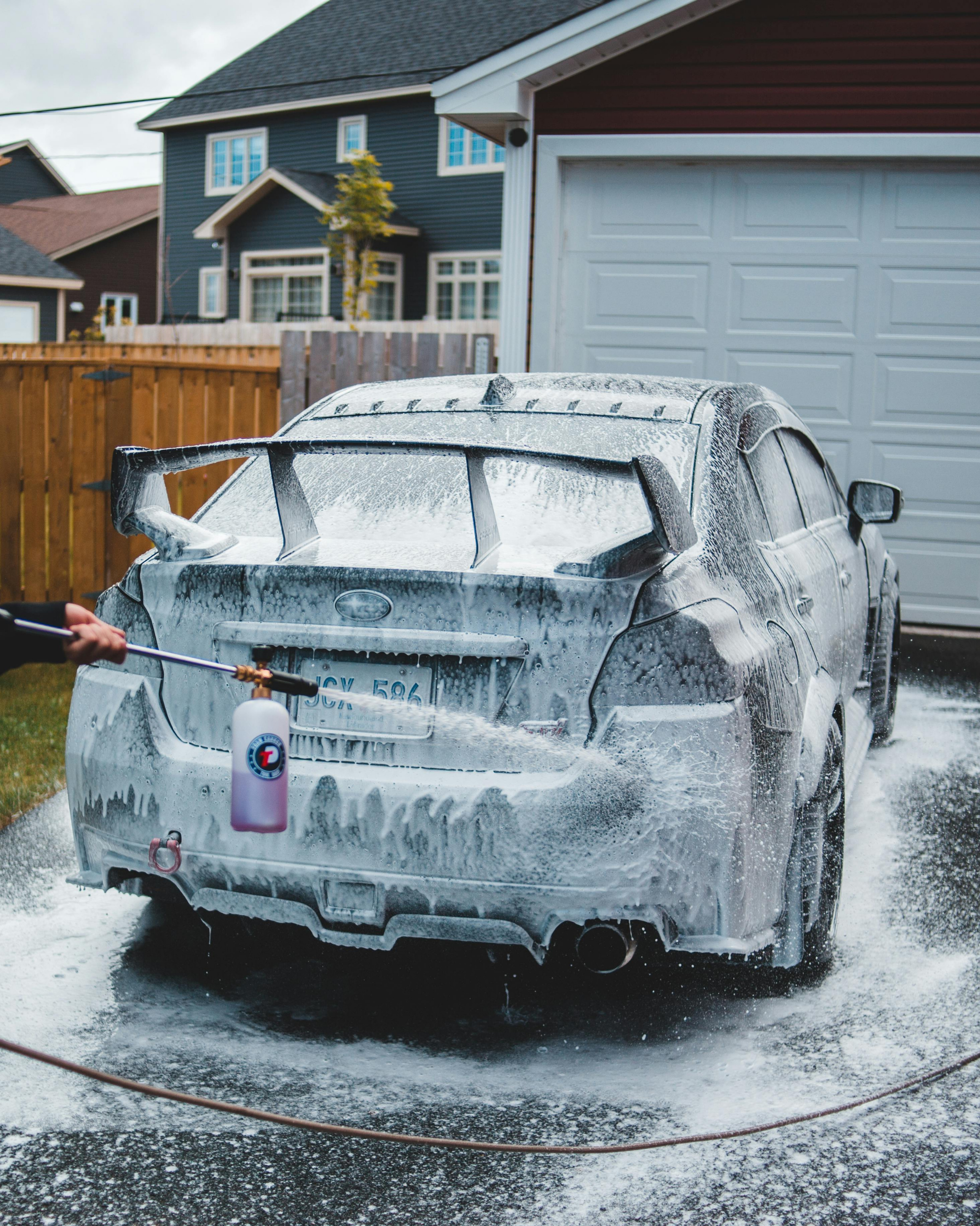 Person washing a gray car with soap suds and foam in a driveway