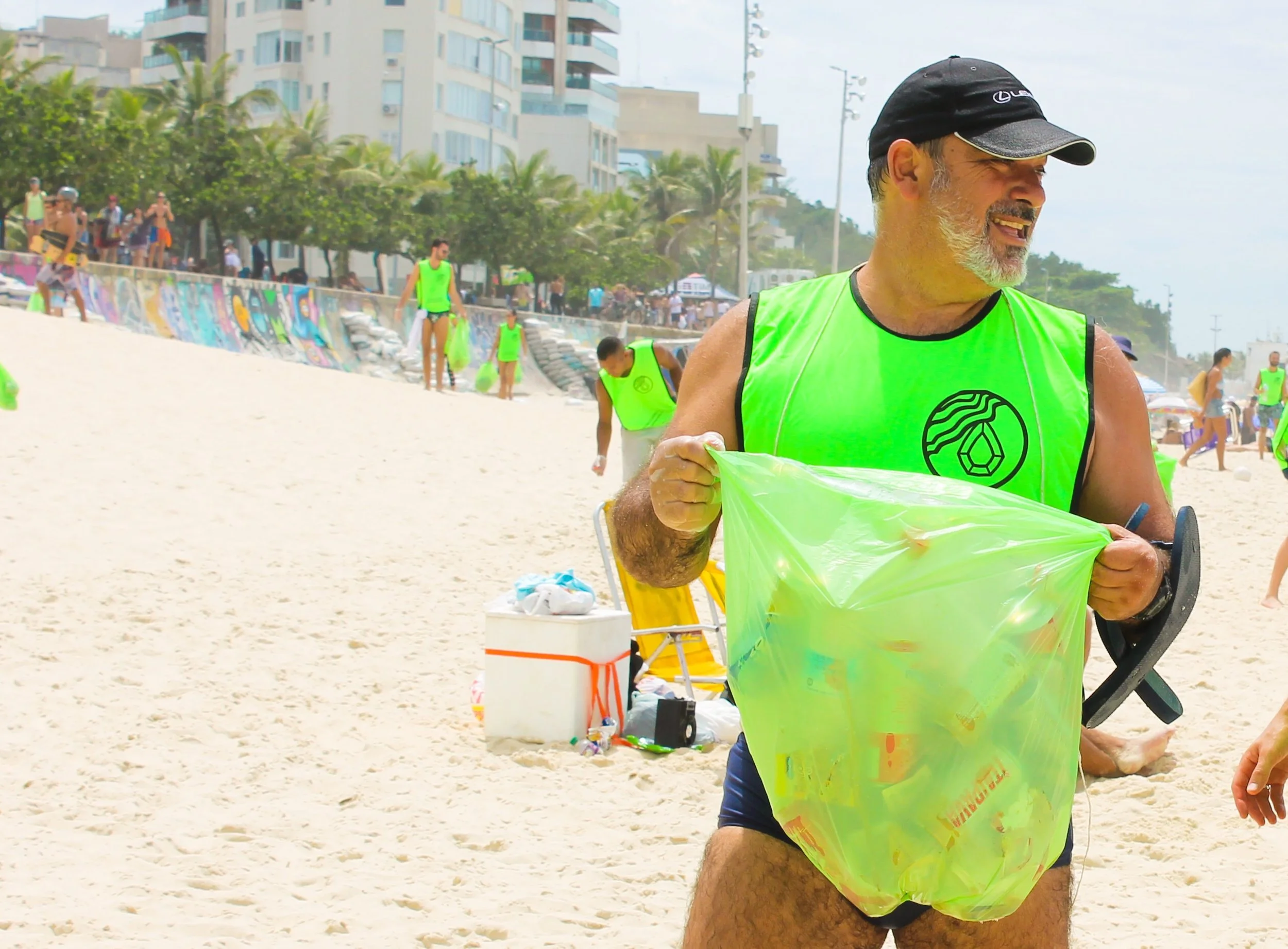 Homem limpando a praia com saco de lixo, usando colete de cor verde neon e boné preto, ao centro da imagem com várias pessoas ao fundo também recolhendo lixo na areia.