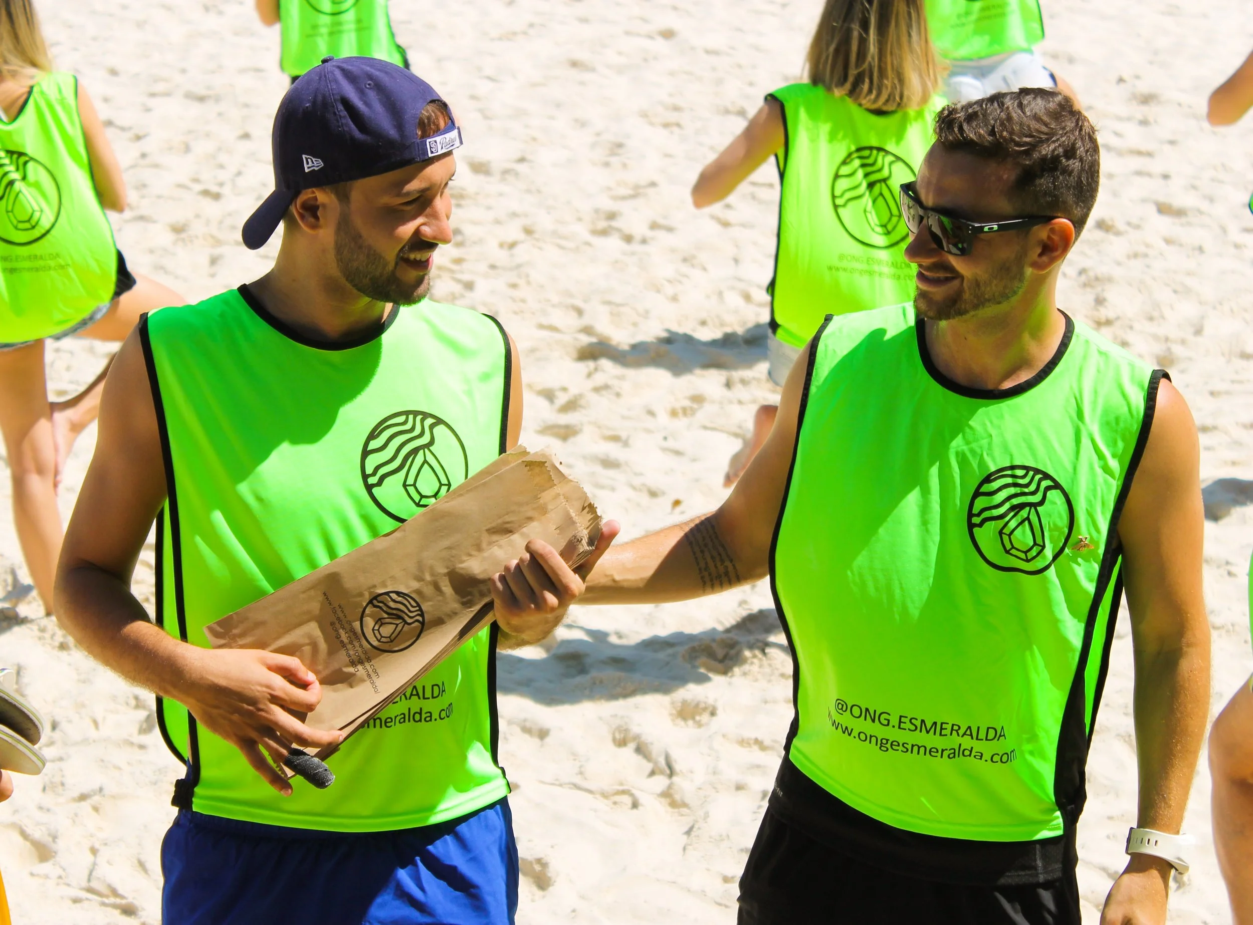 Homens de camiseta laranja na praia, entregando sacola para mulher na areia.