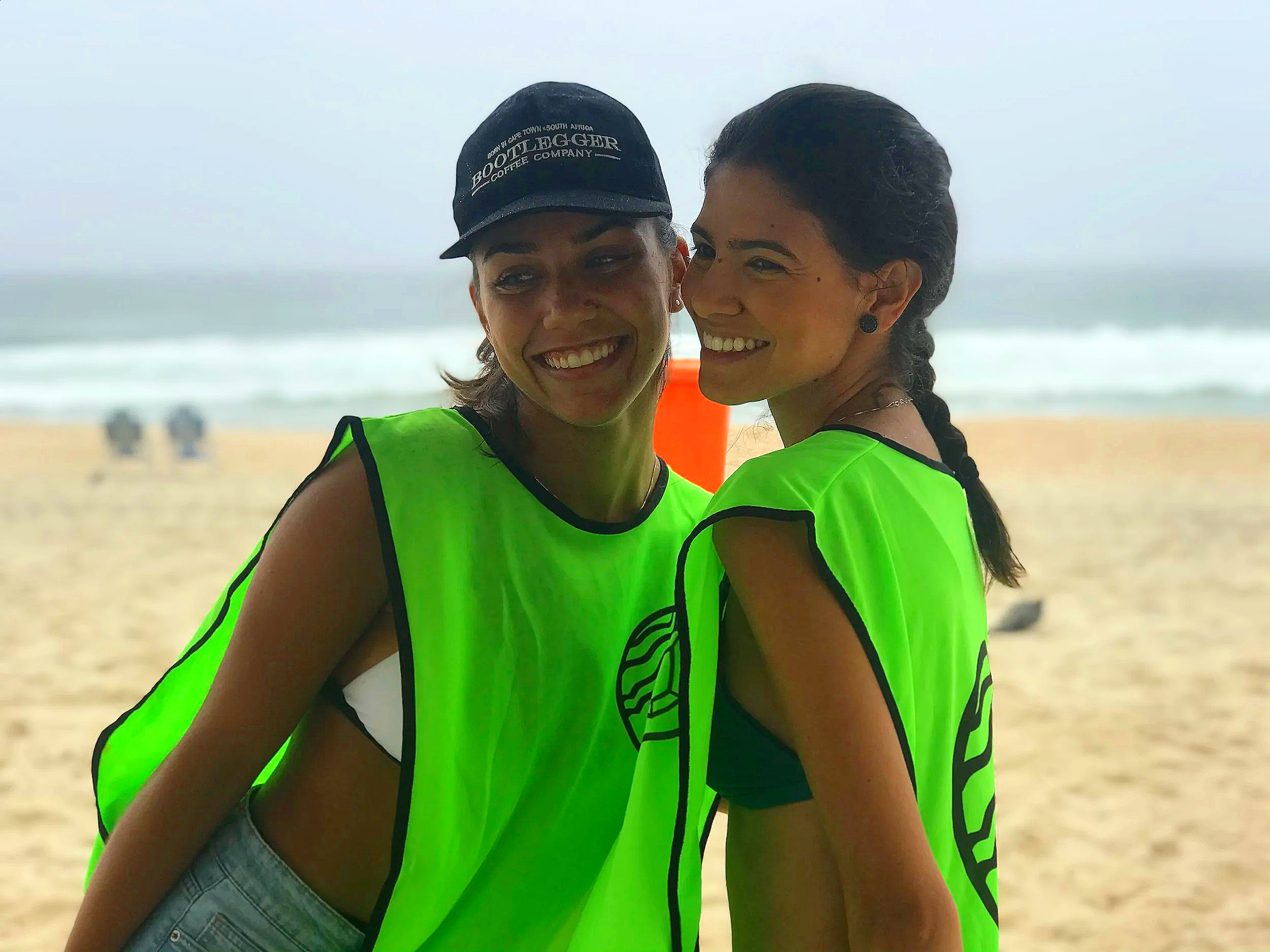 Duas mulheres sorrindo na praia, usando coletes de proteção verdes, ao fundo areia e mar com ondas.