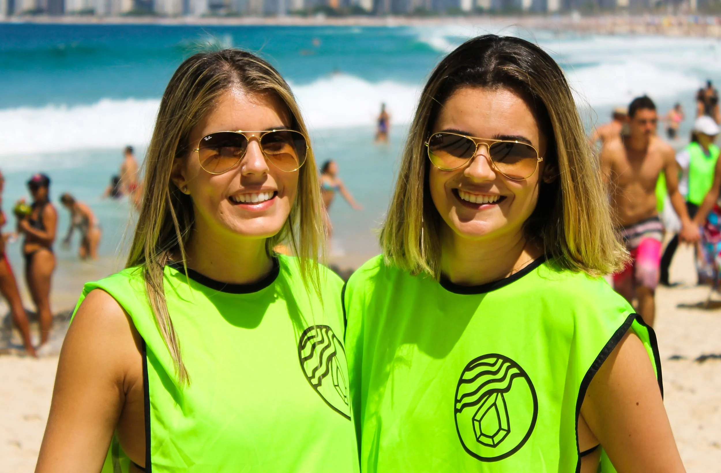 Duas mulheres sorrindo na praia, usando coletes de cor neon e óculos escuros, com o mar e outras pessoas ao fundo, fazendo limpeza de praia