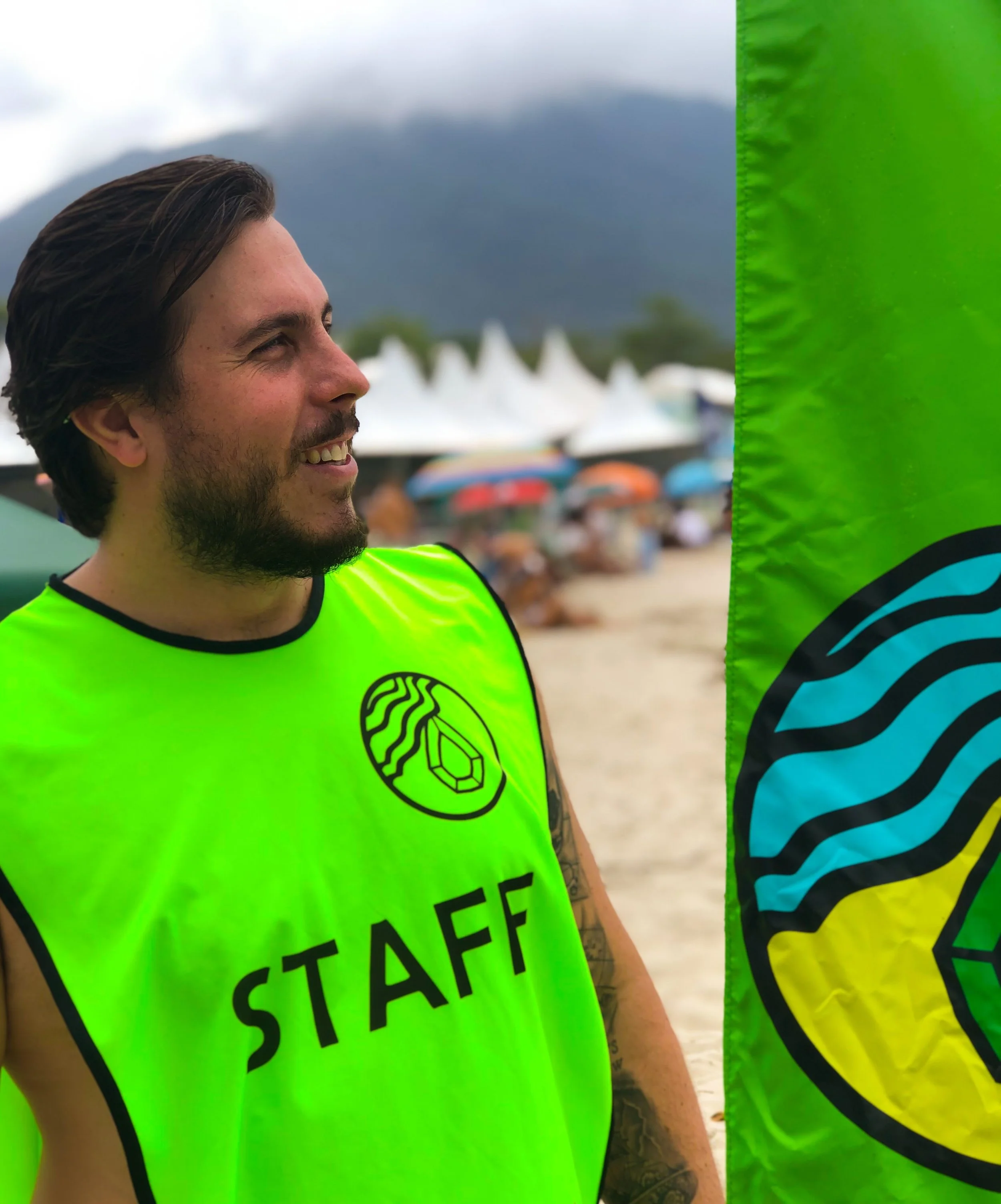 Homem sorrindo usando colete verde com a palavra 'STAFF' e um logotipo, ao lado de uma bandeira com o mesmo logotipo, em uma praia com muitas pessoas e cabanas ao fundo.