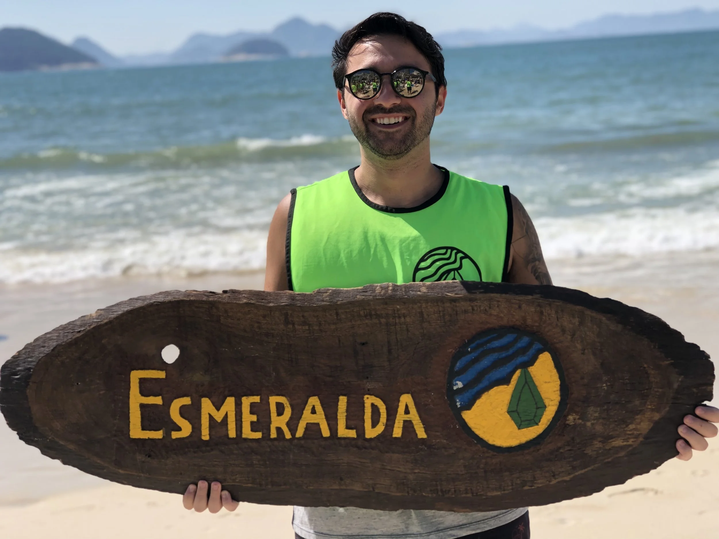 Homem sorridente na praia segurando uma placa de madeira com a palavra 'Esmeralda' e um símbolo colorido de uma pedra preciosa. Voluntário, voluntariado, trabalho voluntário.
