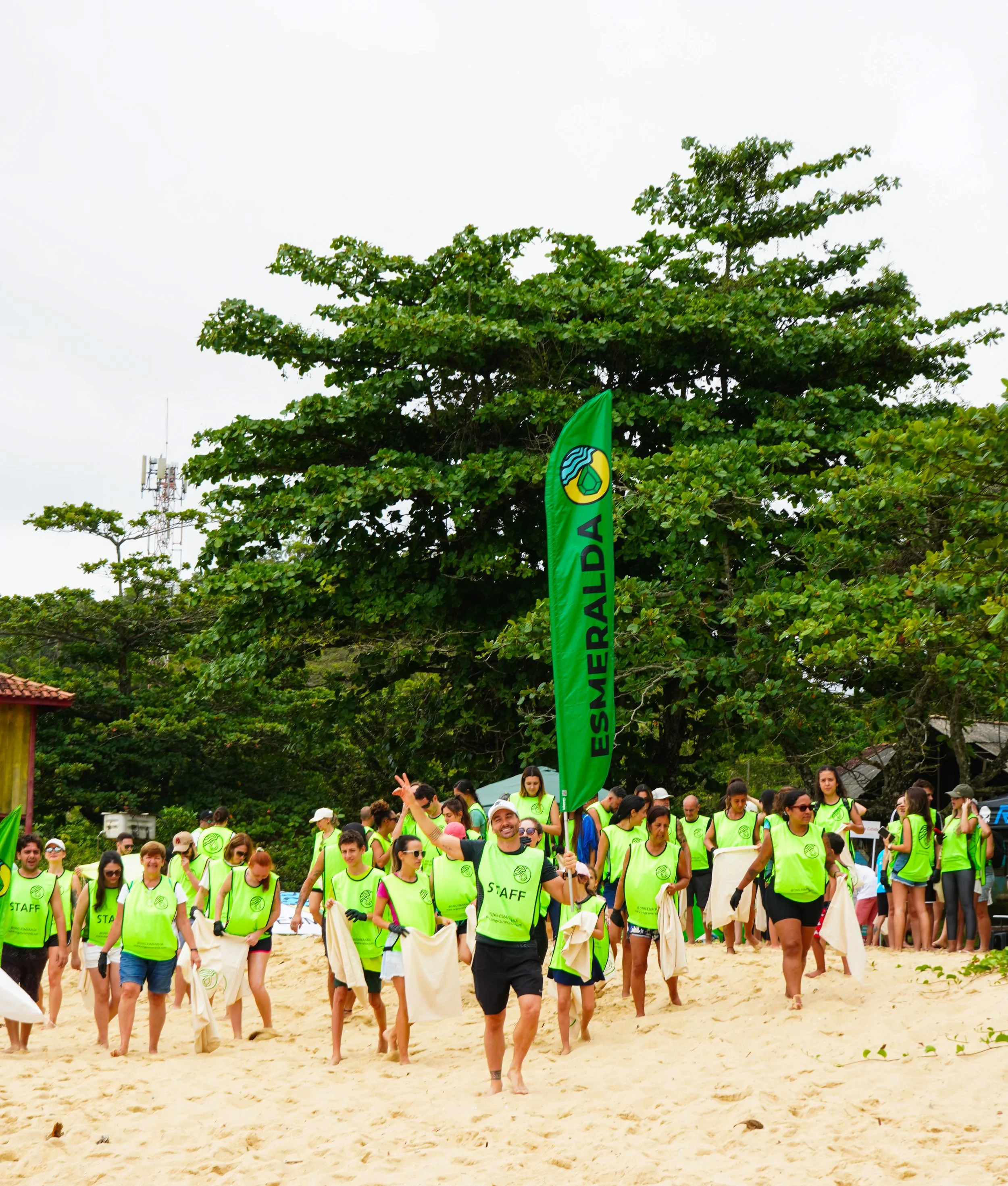 Grupo de pessoas participando de uma atividade na praia, vestindo coletes verdes, com uma bandeira verde que diz 'Esmeralda' e um logo verde, ao fundo árvores e céu nublado. Limpeza de praia, conscientização e consciência ambiental, solidariedade