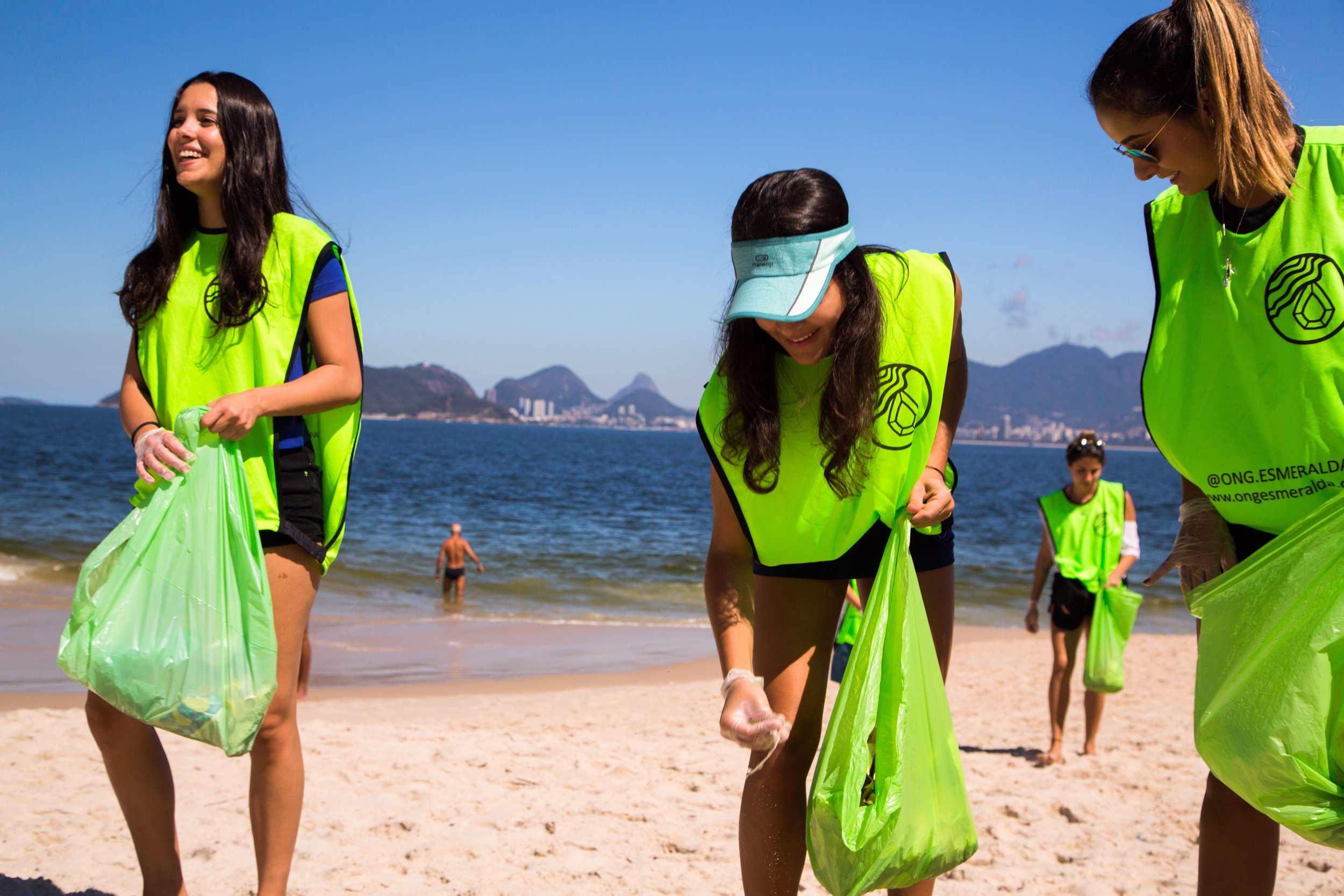 Grupo de pessoas, mulheres jovens, participando de limpeza na praia durante um dia ensolarado, vestindo coletes de neon, segurando sacos de lixo verdes, com o mar e montanhas ao fundo.