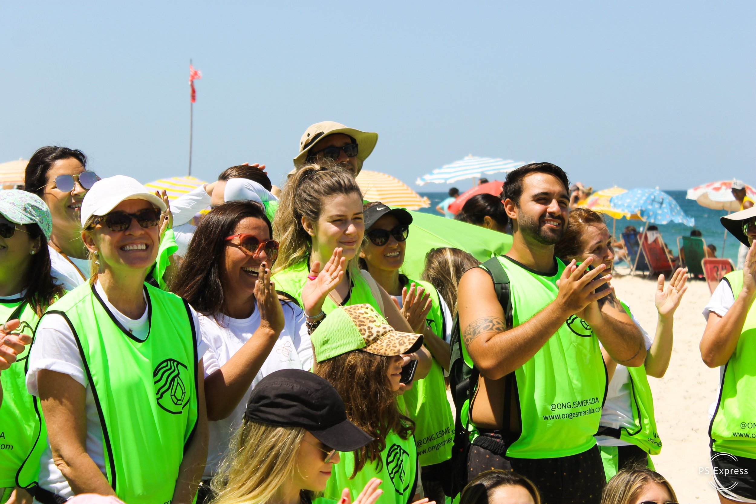 Grupo de pessoas na praia usando coletes verdes, sorrindo e assistindo a um evento na areia com guarda-sóis ao fundo.