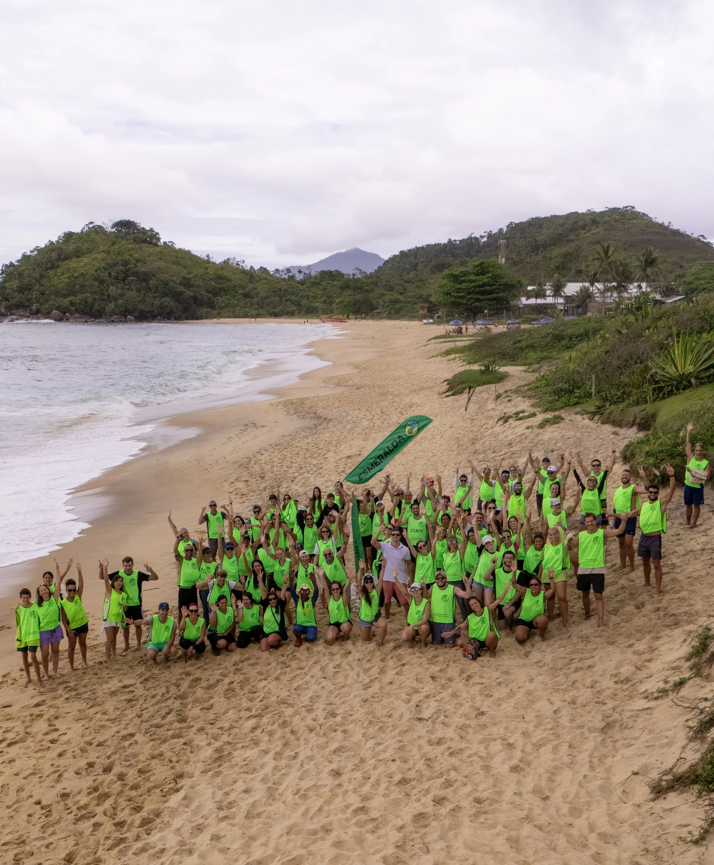 Grupo de pessoas usando coletes verdes na praia com vegetação e colinas ao fundo