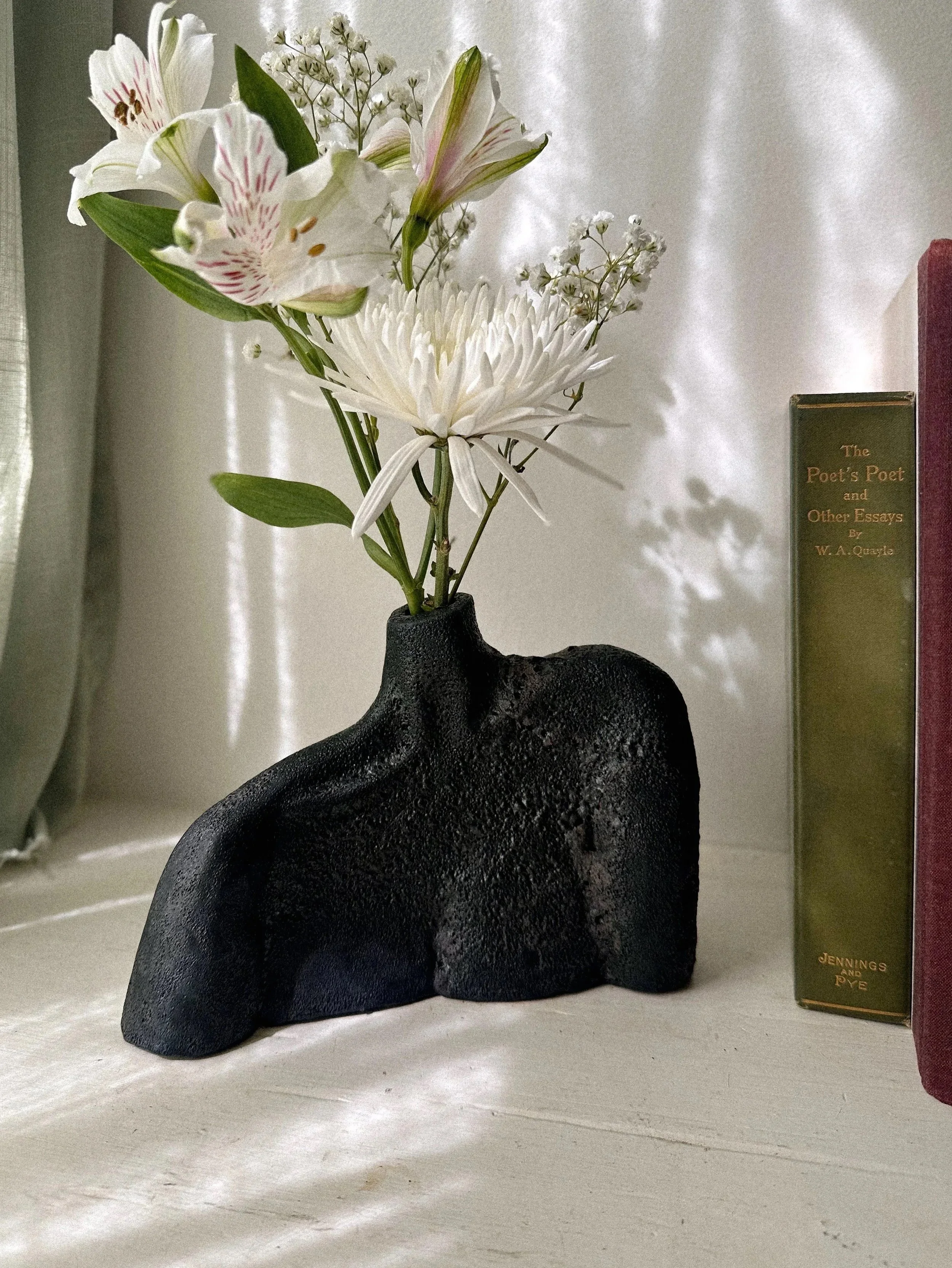 A black textured ceramic vase with a unique shape holds a bouquet of white and pink flowers. The vase is placed on a cream-colored surface next to a green book titled "The Poet's Poet and Other Essays" by W. A. Quayle and a red book. Light and shadow create soft reflections on a white curtain in the background.