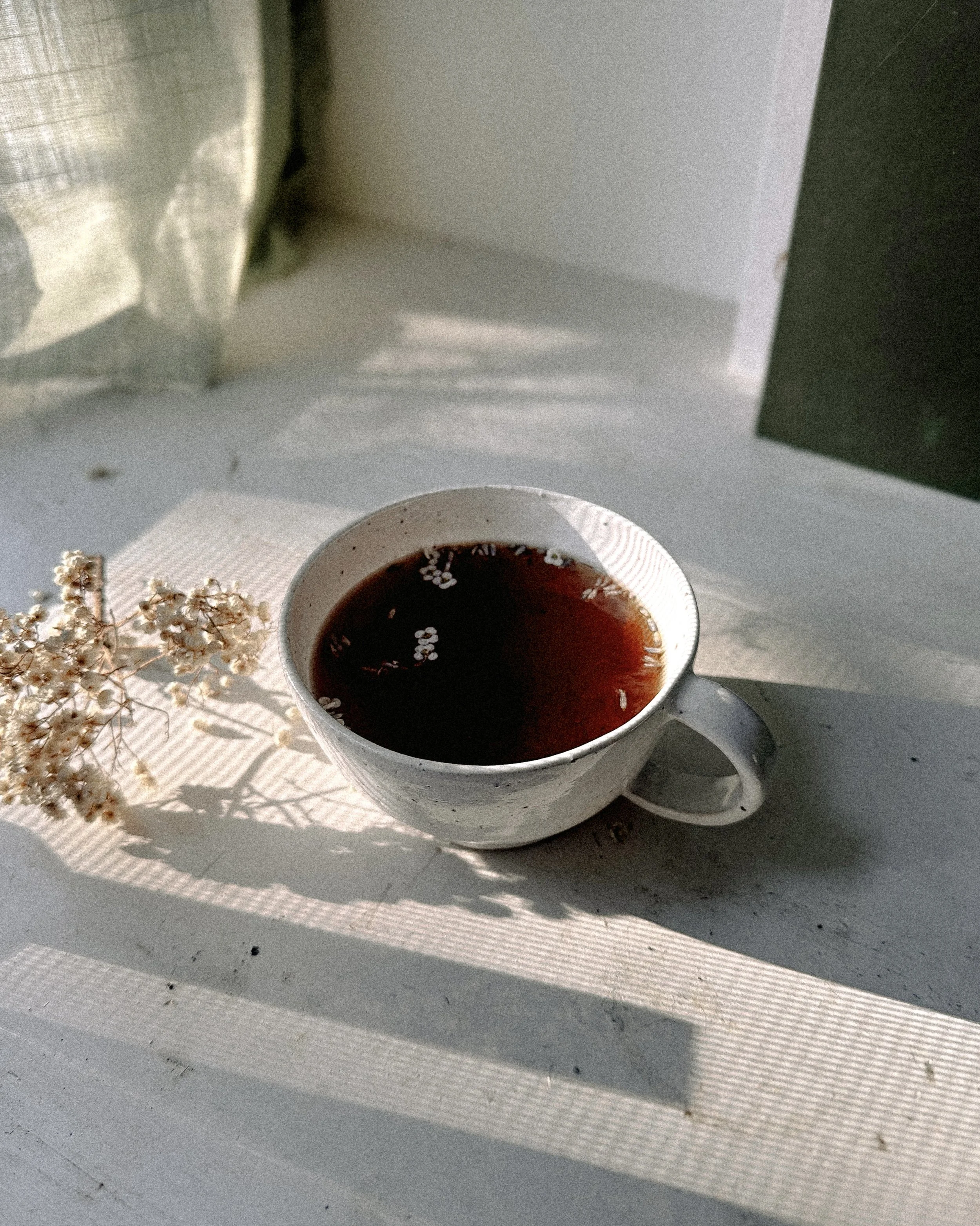 A white ceramic cup filled with black tea placed on a white surface, with shadows cast by sunlight, next to dried flowers and a partially visible curtain in the background.