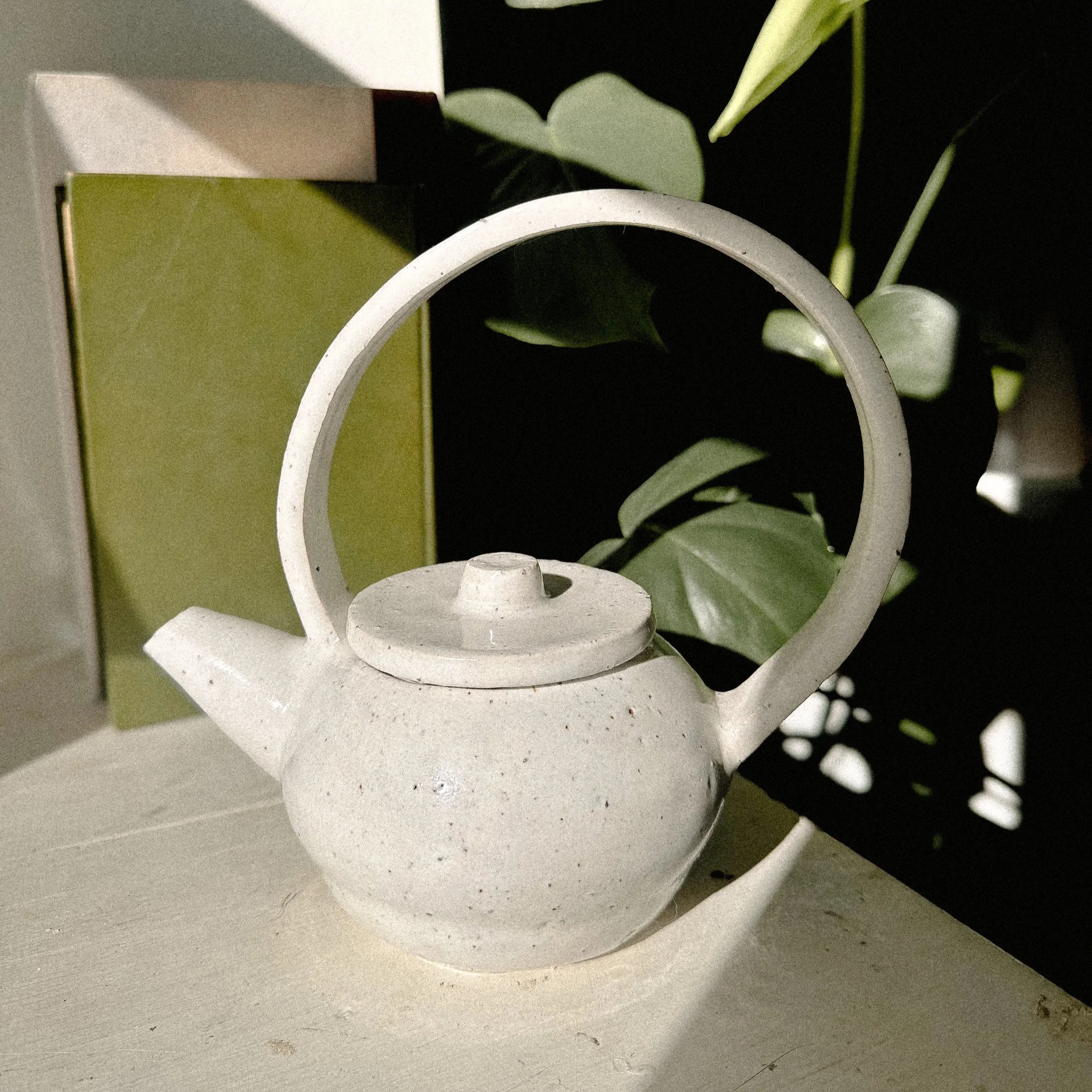 White ceramic teapot with a large curved handle, placed on a light surface, with green leaves and a beige box in the background.