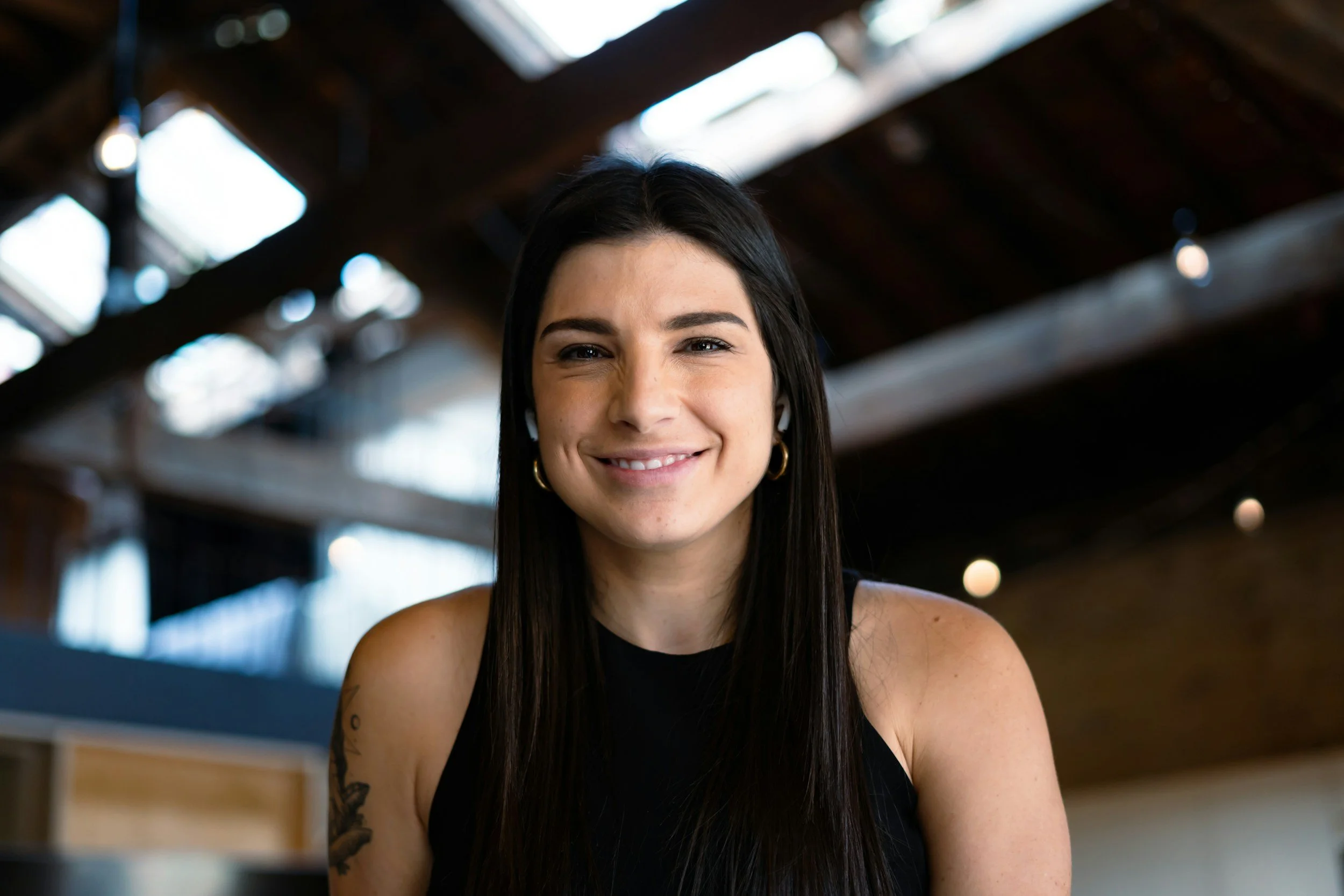 Smiling young woman with long dark hair, wearing a black sleeveless top, sitting indoors with a high ceiling and wooden beams in the background.