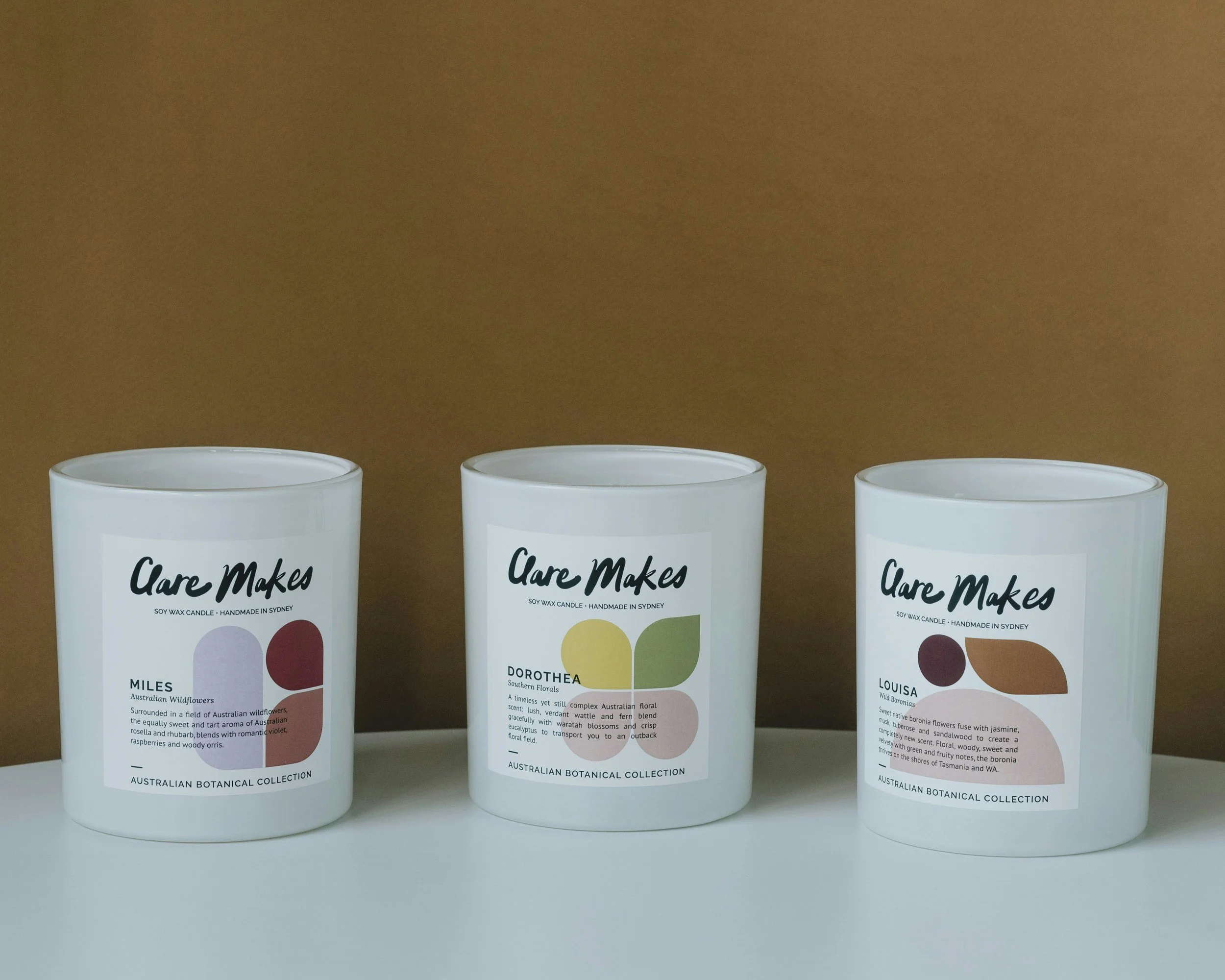 Three white candles labeled 'Care Makes' from an Australian botanical collection, each with different botanical names and colorful abstract designs, placed on a white surface against a brown background.