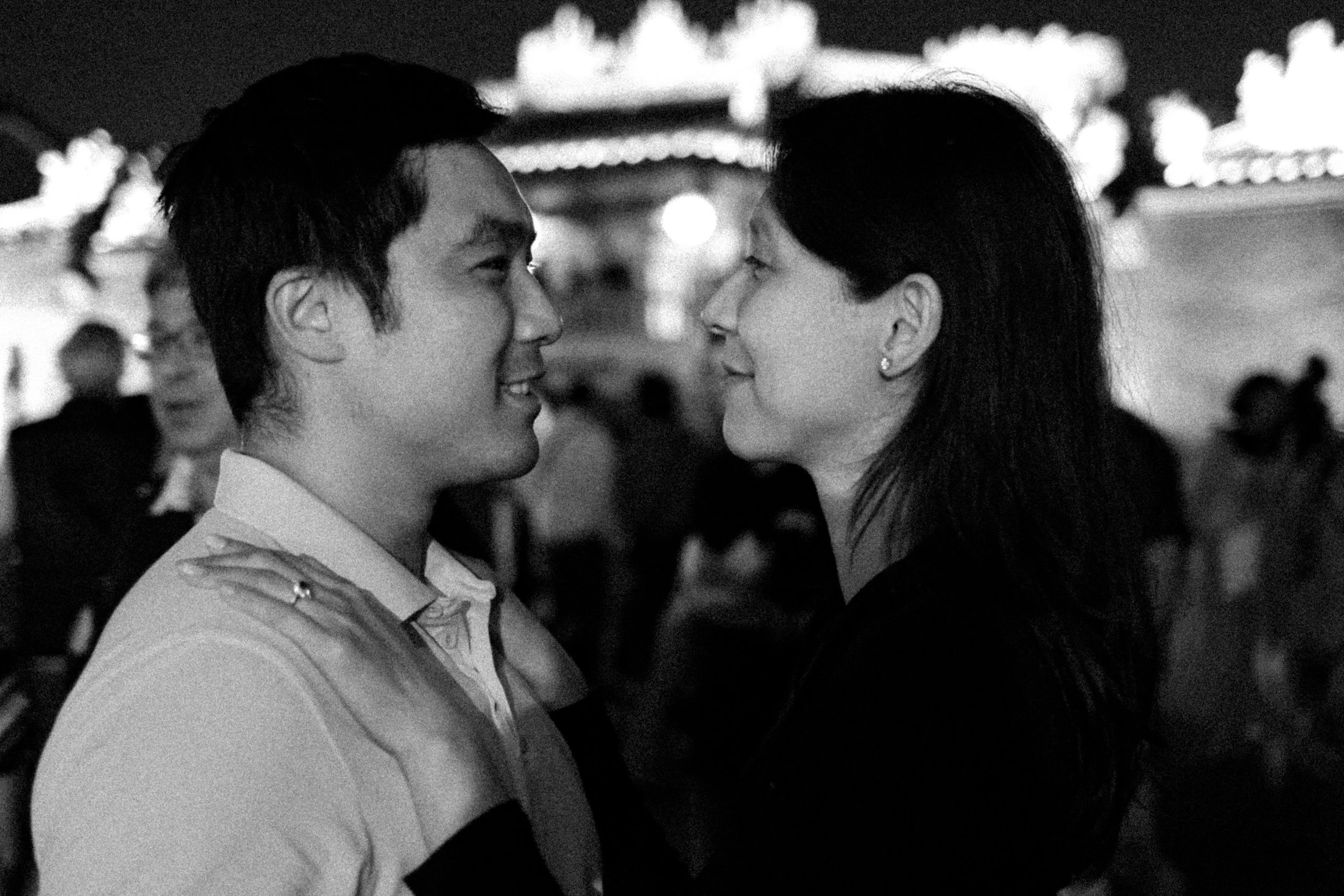 A black and white photo of a couple standing close to each other, smiling, at an outdoor event during nighttime with blurred lights in the background.