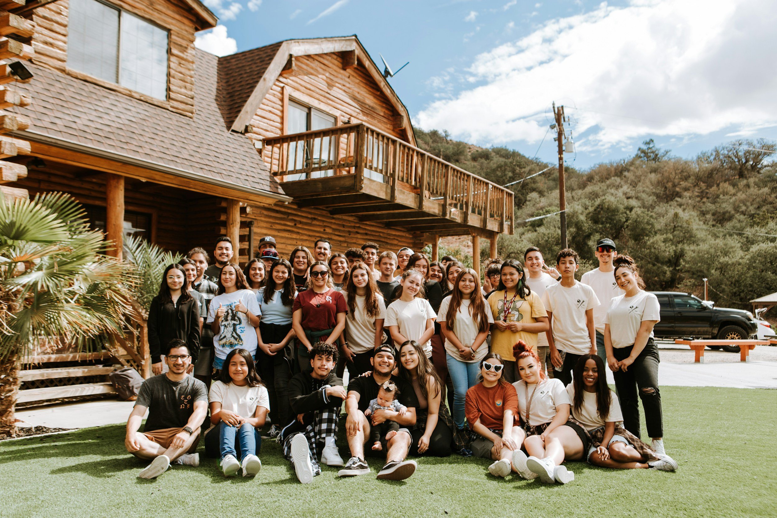 A large group of young people posing for a photo outside a wooden house on a sunny day, with trees and mountains in the background.