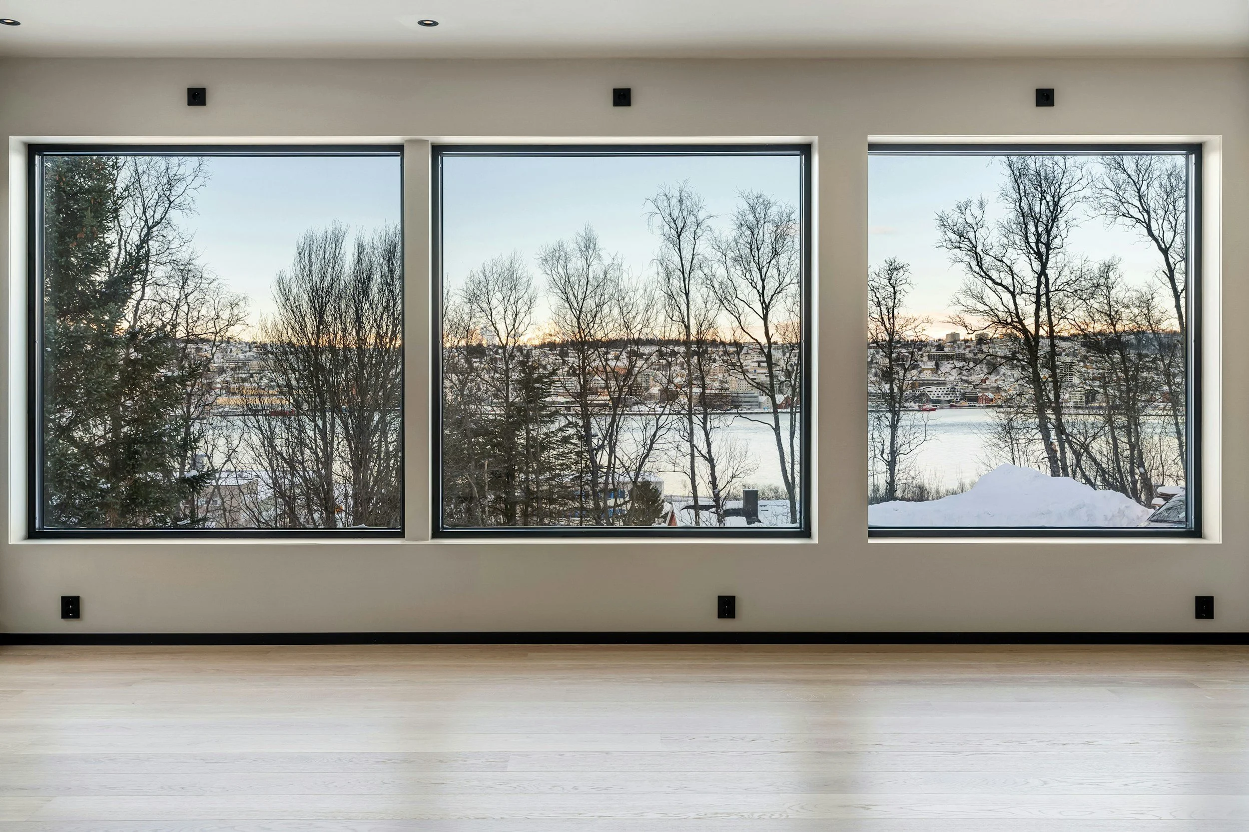 Empty room with large three-pane windows showing a snowy winter landscape with trees and water outside.