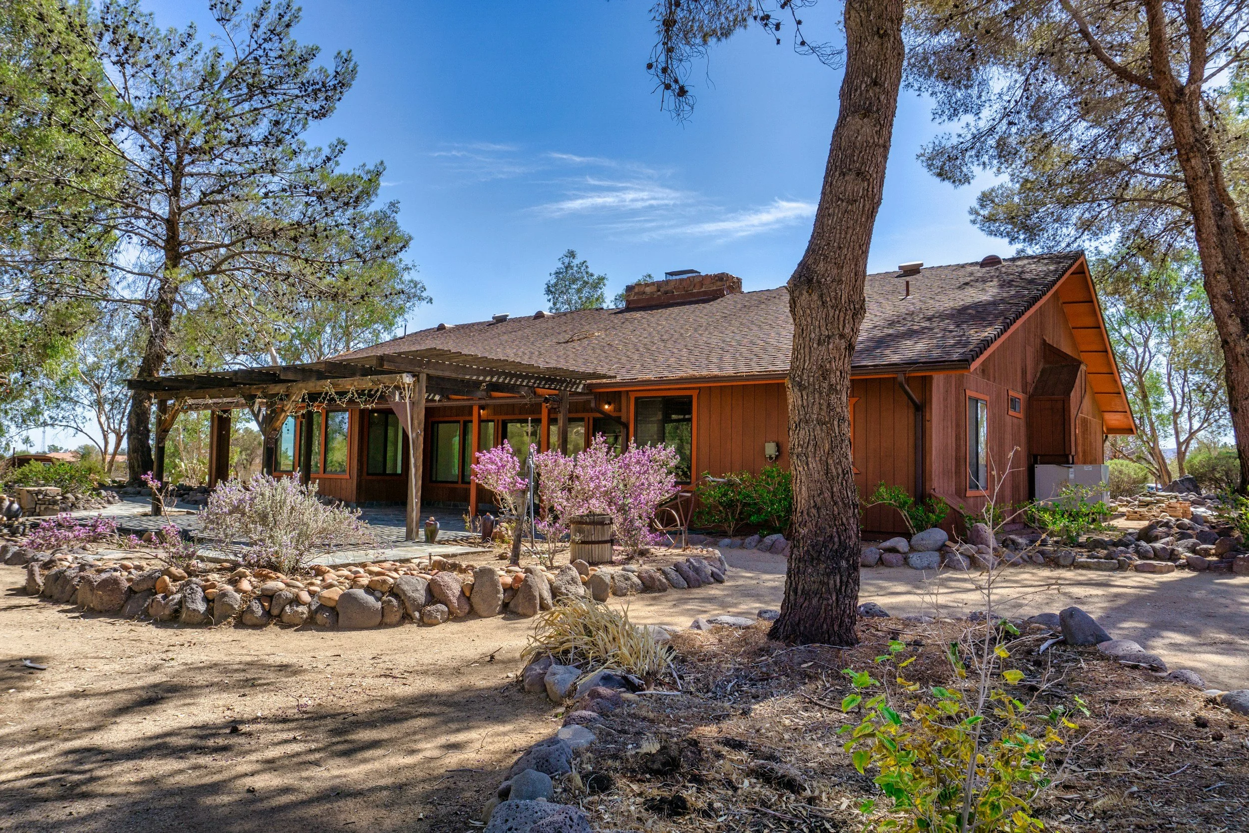 A single-story wooden house with a shingled roof and large windows, surrounded by trees, desert plants, and rocks in a dry landscape under a blue sky.