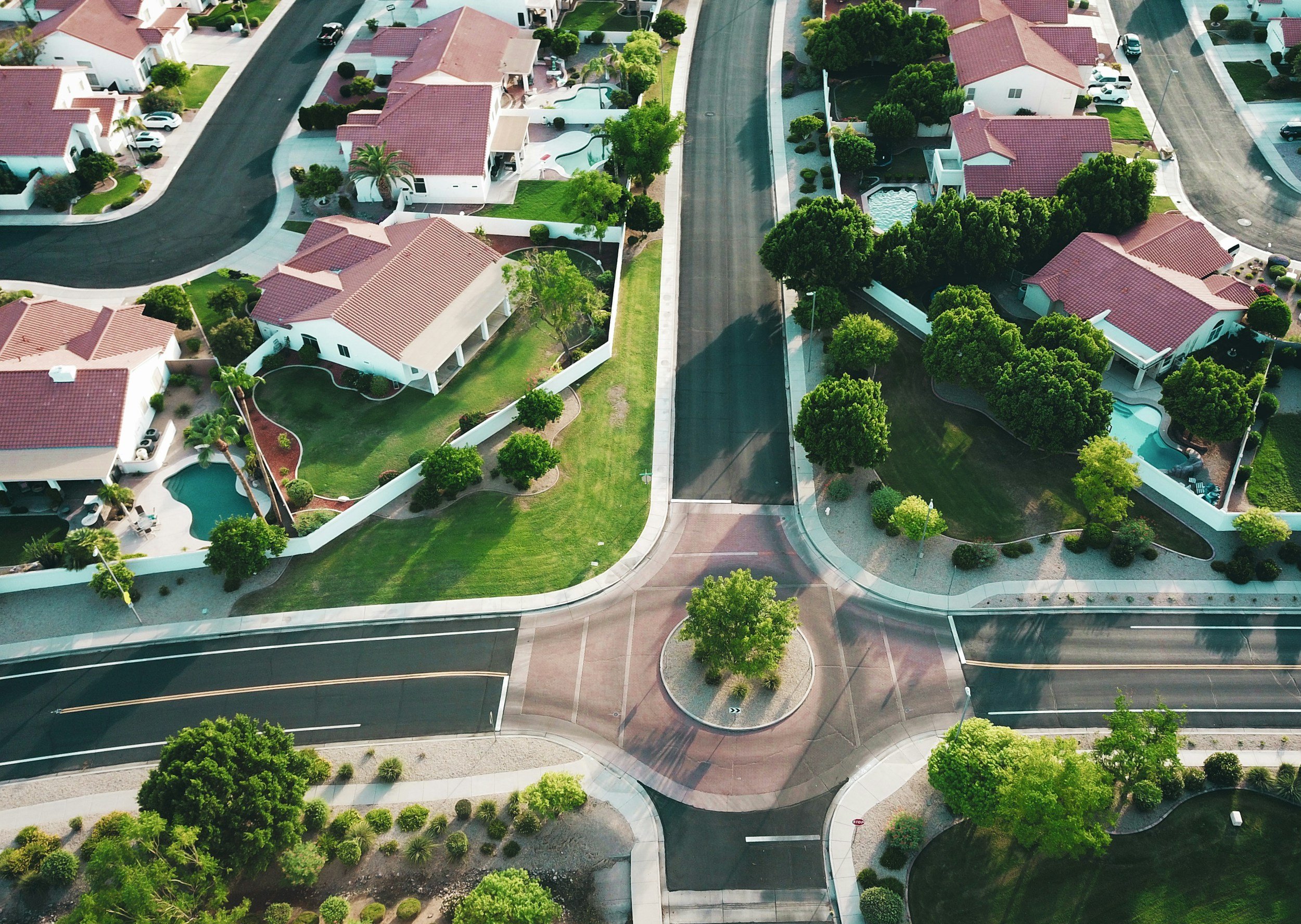 An aerial view of a suburban neighborhood with single-family homes, manicured lawns, swimming pools, and trees. There are quiet streets, a landscaped roundabout with a tree, and well-maintained yards.