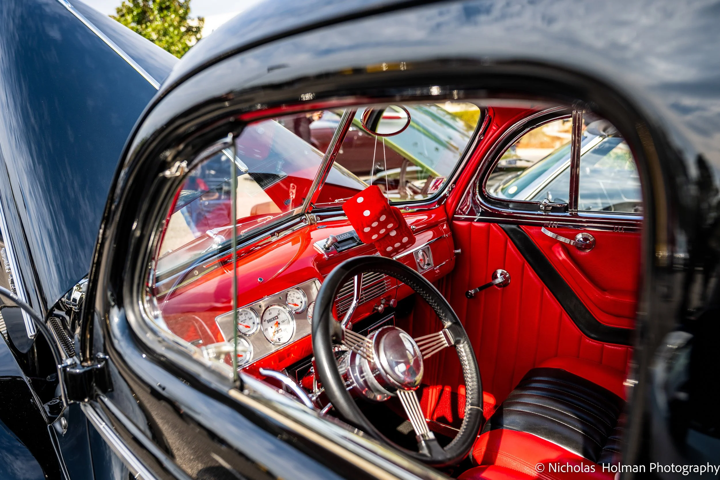 Interior view of a vintage car with red and black interior, dashboard with gauges, black steering wheel, and red fuzzy dice hanging from the mirror.