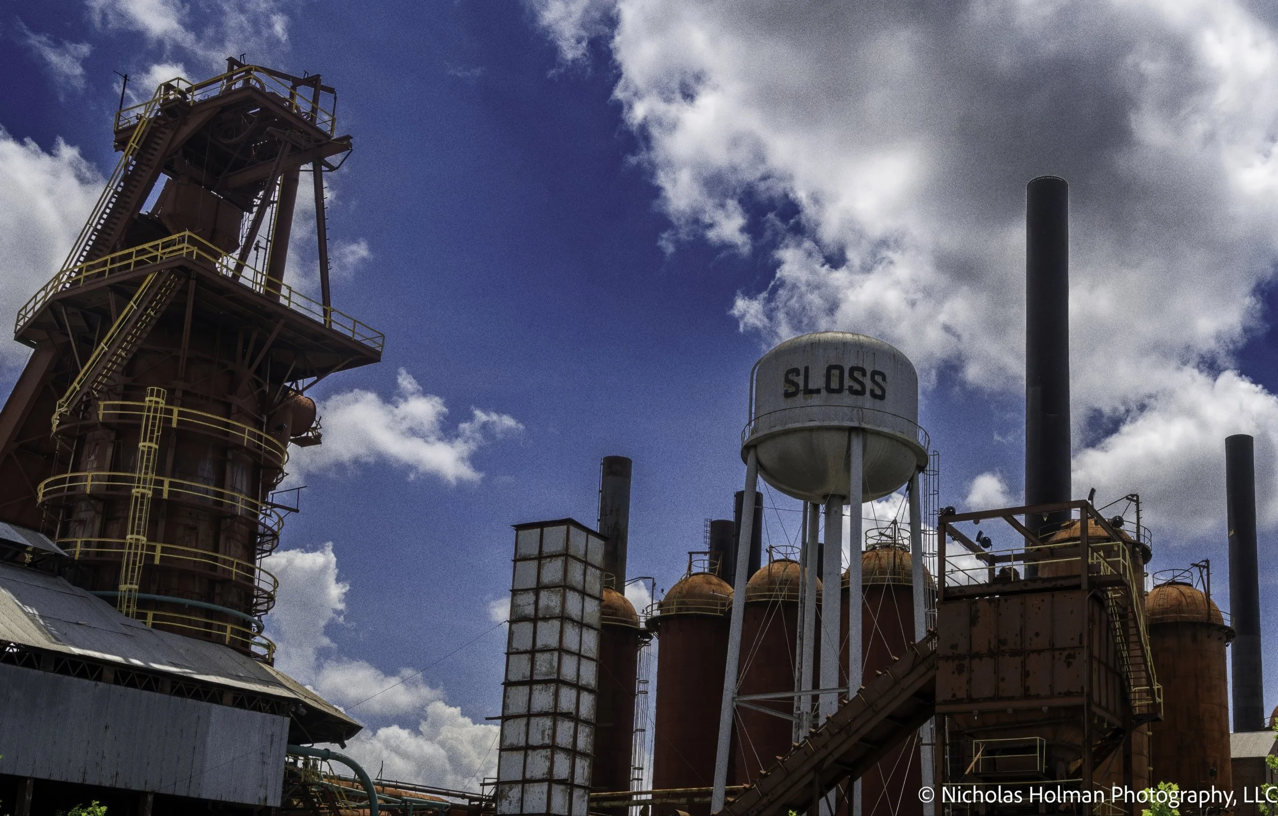 Industrial plant with red metal structures, yellow railings, multiple tanks, including a water tower labeled 'SLOSS,' and several smokestacks against a partly cloudy sky.