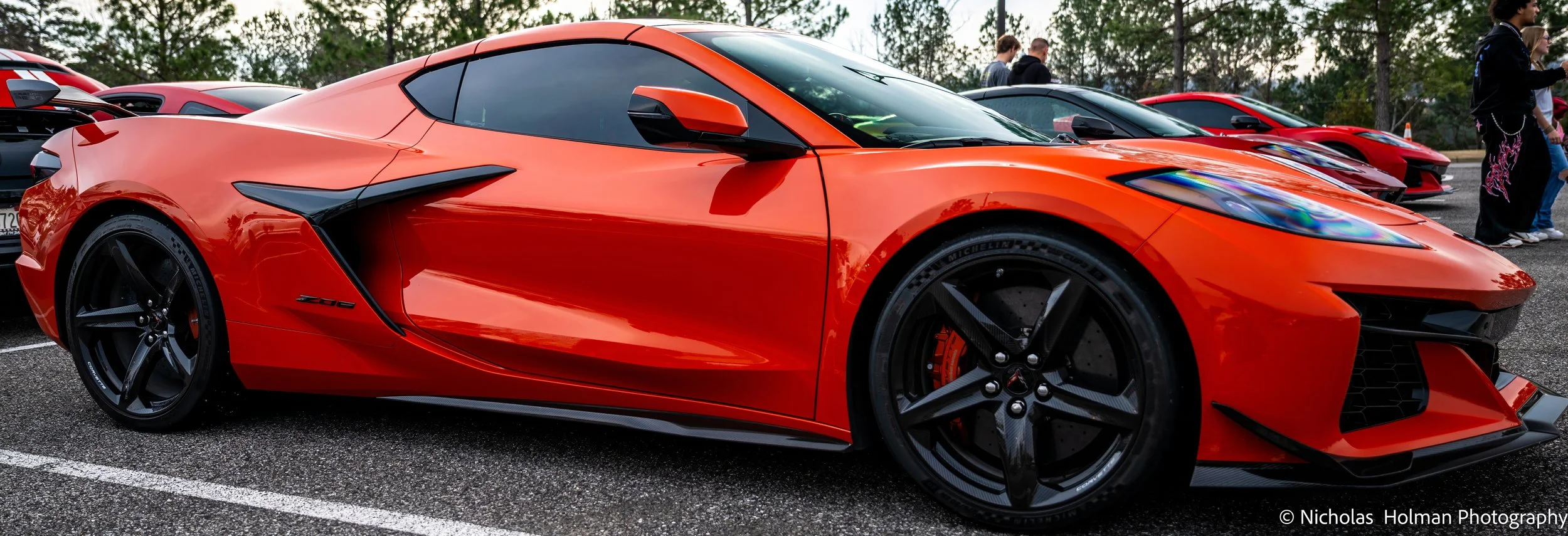 Red Chevrolet Corvette Z06 sports car parked in a lot during daytime with other cars and people in the background.
