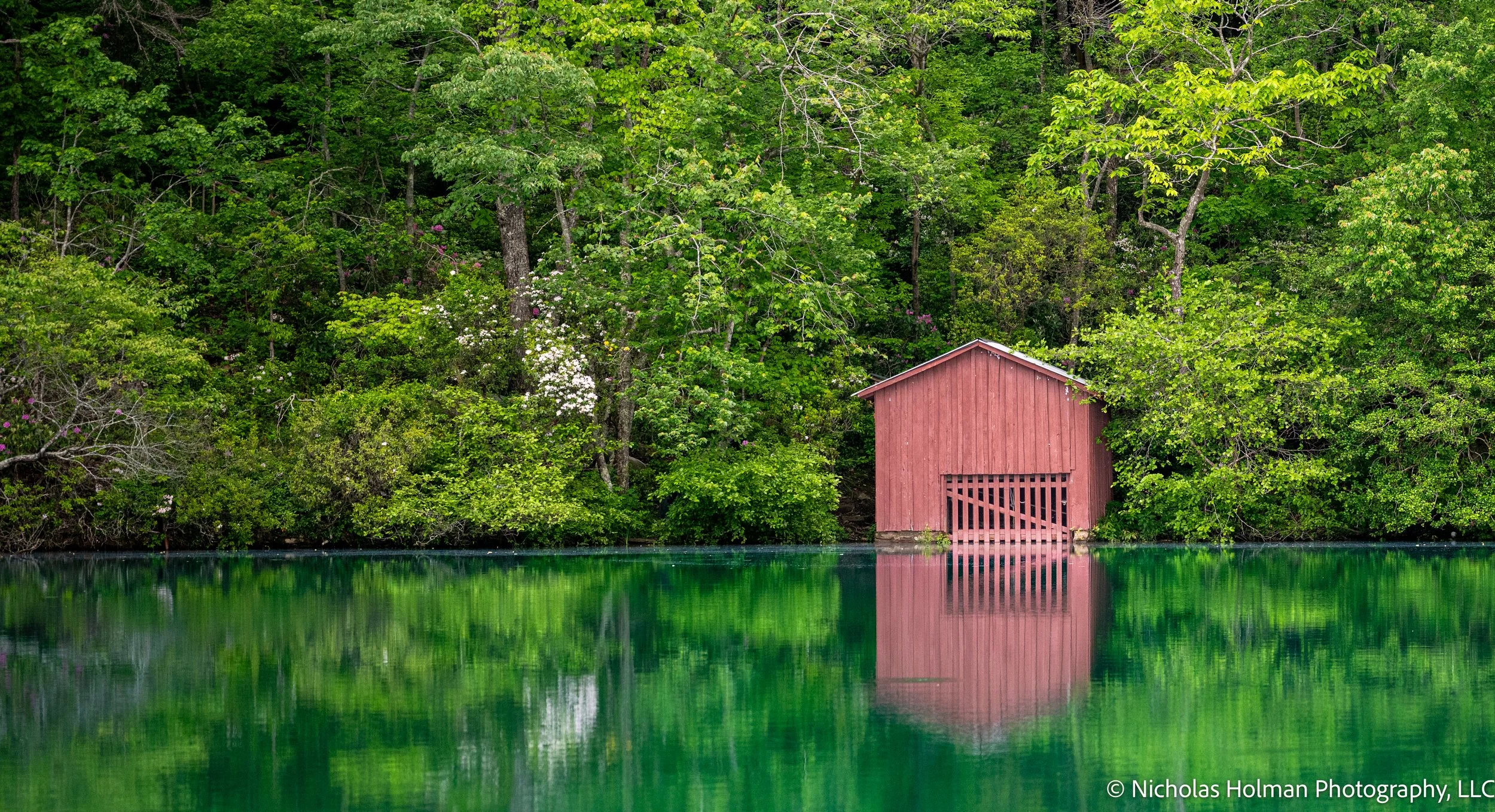 A red boathouse by a lake, surrounded by dense green trees and foliage, with their reflection in the water.