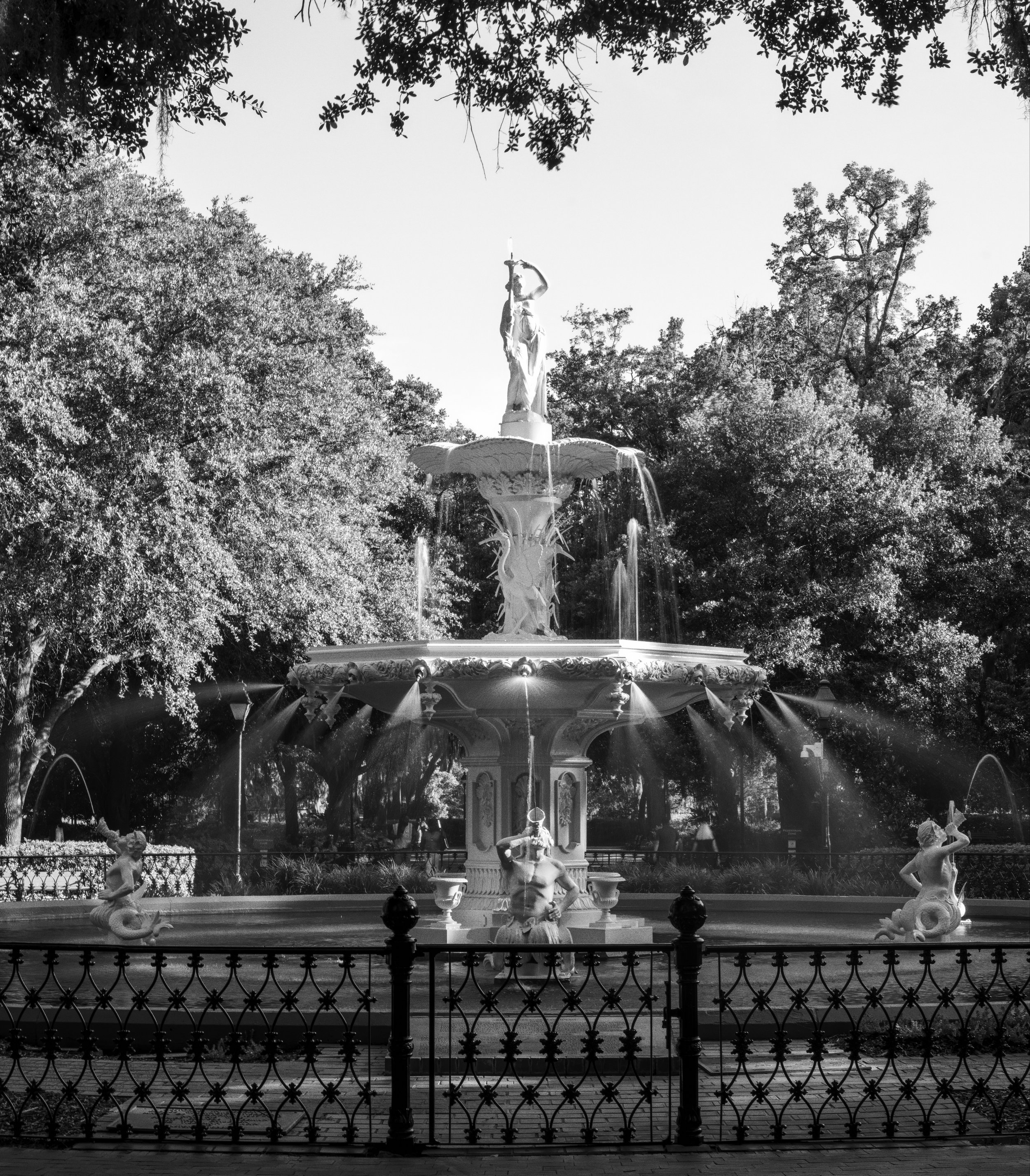 Black and white image of a decorative fountain with sculptures of mermaids around the base, surrounded by a fence, with trees in the background and a clear sky.