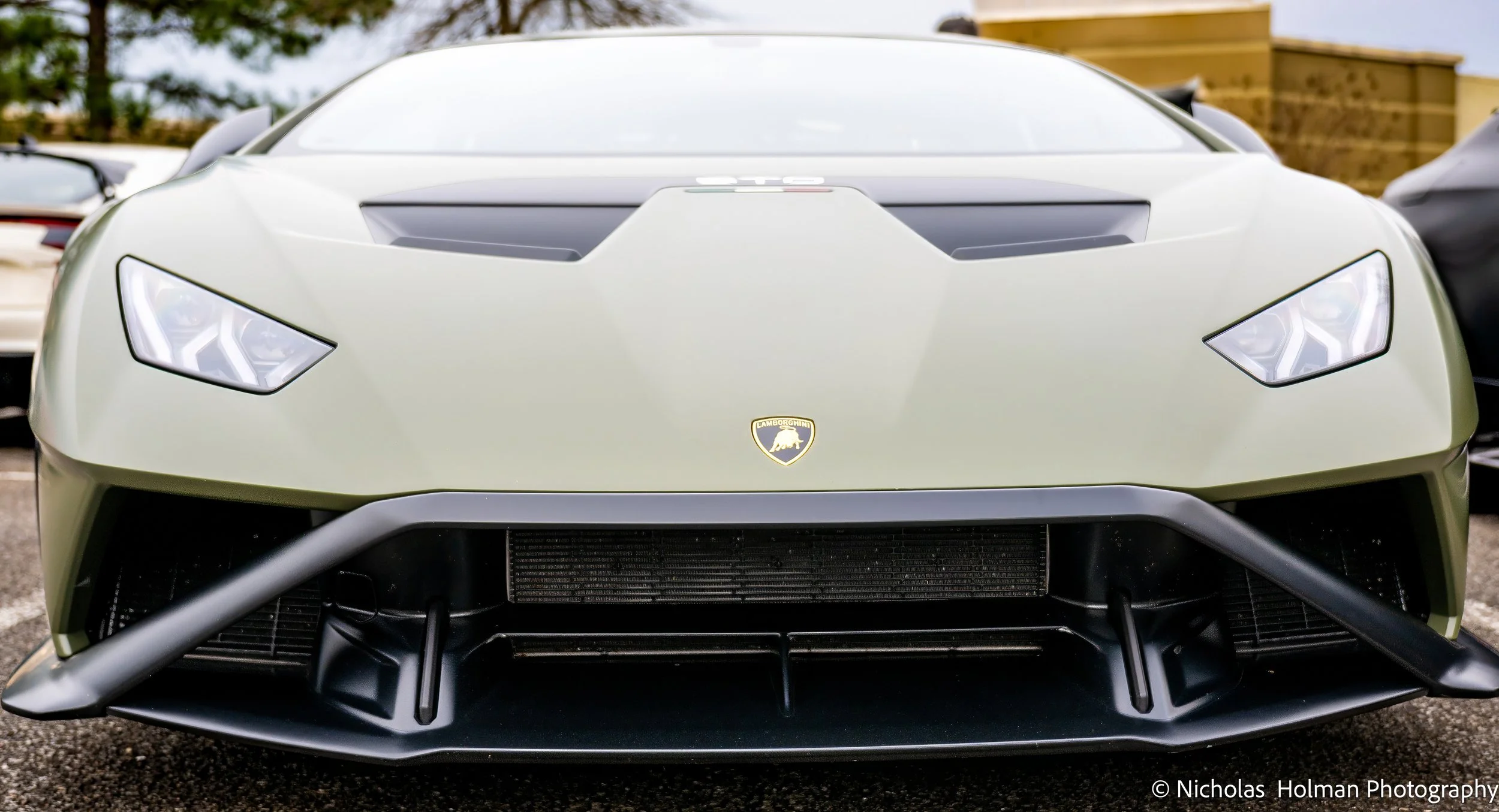 Front view of a Lamborghini Huracan racing car in matte green finish, with a Lamborghini emblem on the hood, aerodynamic splitter, and sharp LED headlights.
