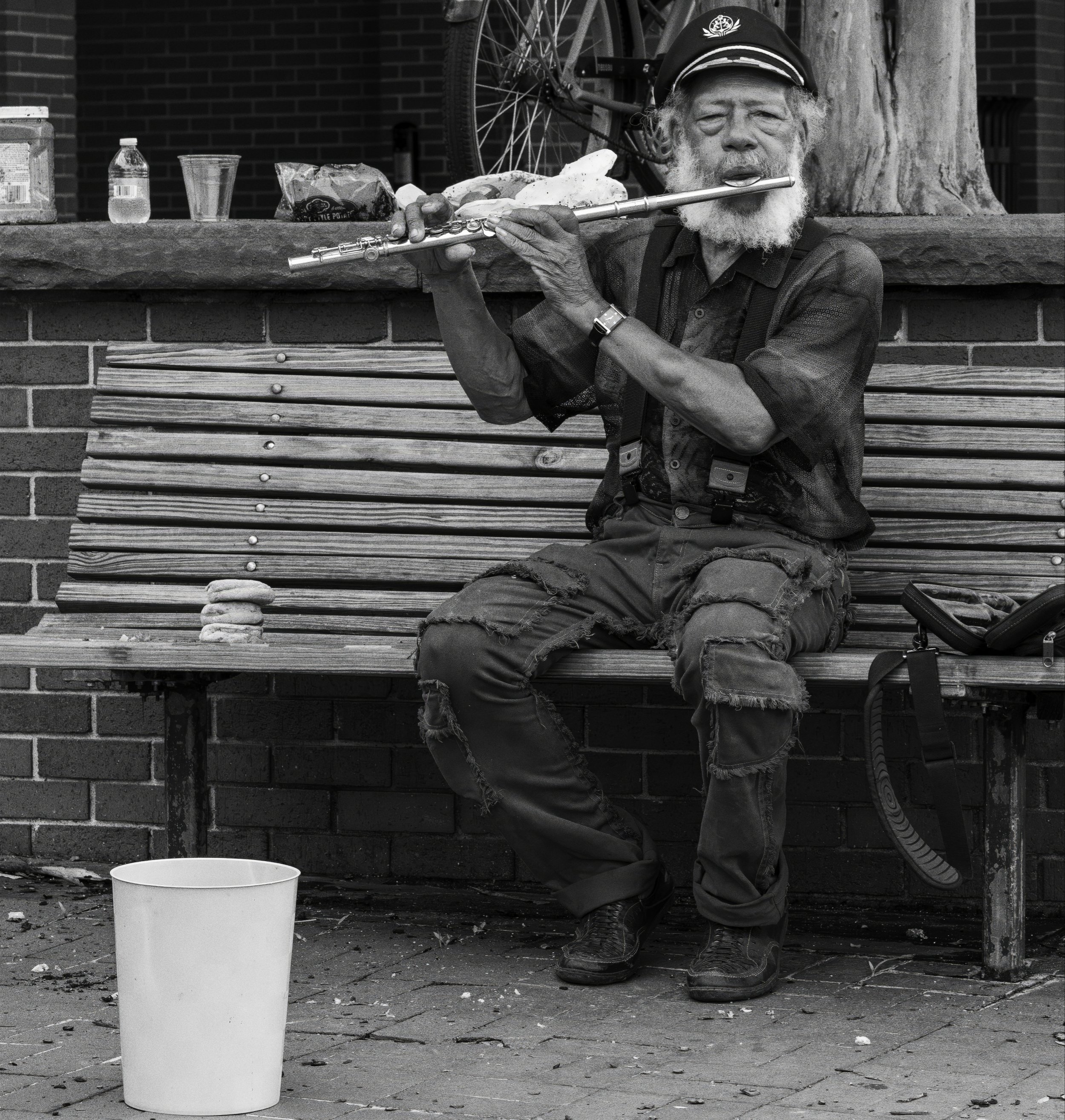 An elderly man with a beard, wearing a cap, sitting on a park bench, playing a flute, with a bicycle, a water bottle, a cup, and food items on the bench behind him, and a white bucket on the ground in front of him.