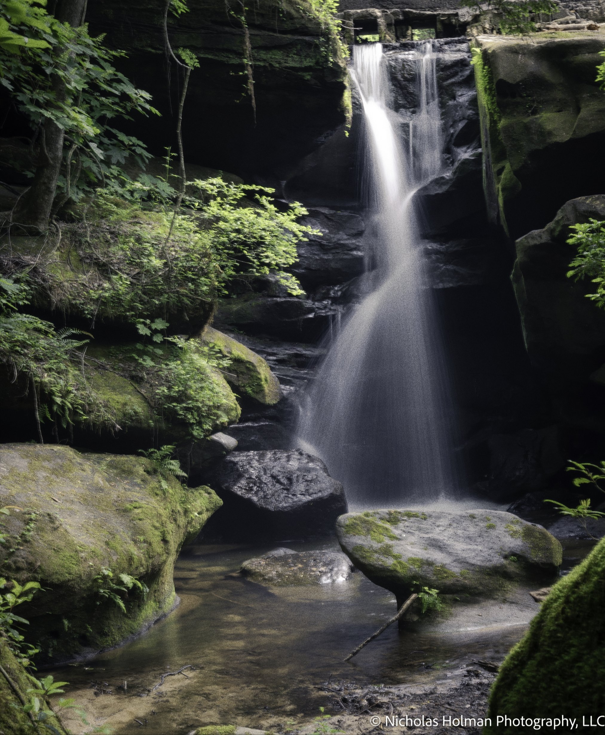 A small waterfall cascading into a stream surrounded by moss-covered rocks and lush green trees in a forest.