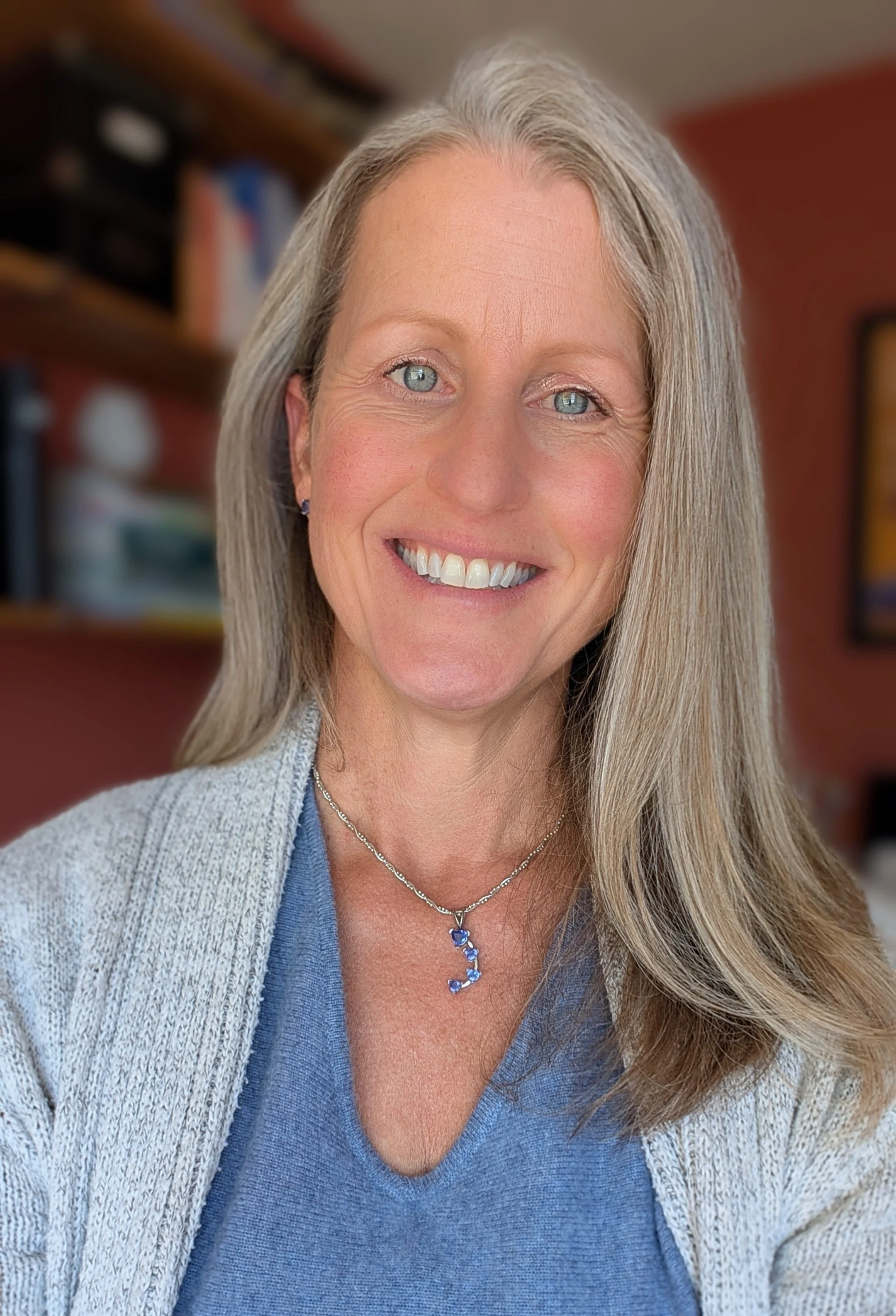 A smiling woman with long blonde hair, wearing a light gray cardigan over a blue top and a necklace with a fox pendant, in a room with a blurred bookshelf and wall art in the background.