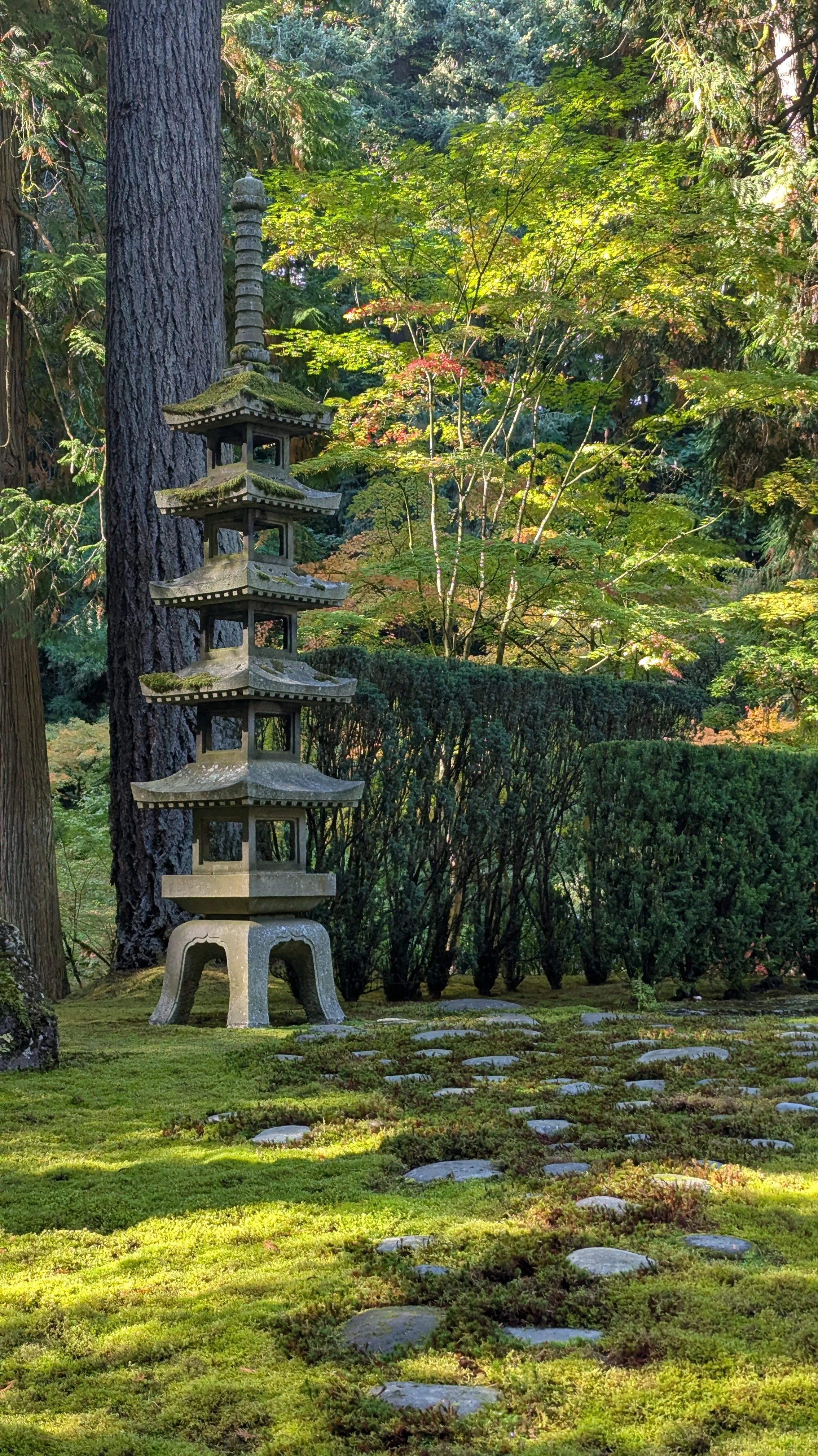 A stone Japanese pagoda sculpture stands in a lush garden surrounded by green moss, shrubs, and trees with sunlight filtering through the leaves.