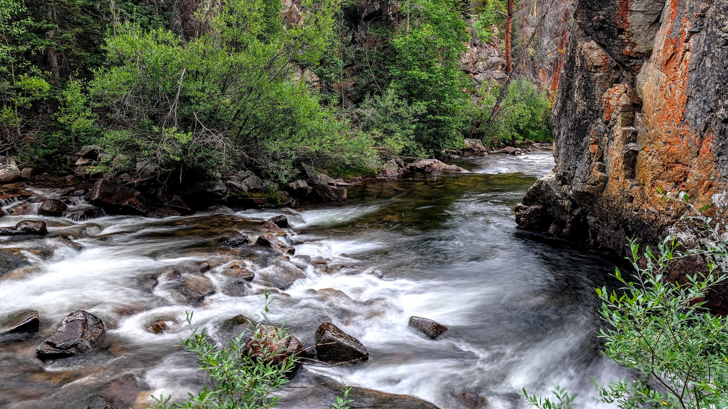 A flowing mountain river with rocks and boulders surrounded by green trees and a rocky cliff.