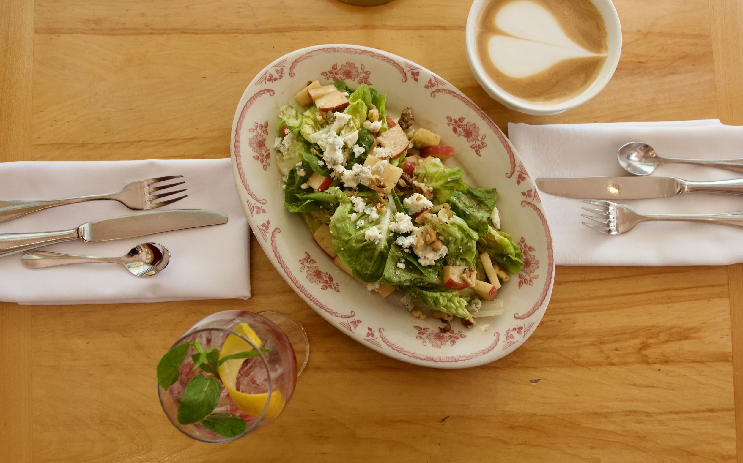 A top-down view of a dining table with a plate of salad, a glass of water with lemon and mint, a cup of coffee, and cutlery on napkins.