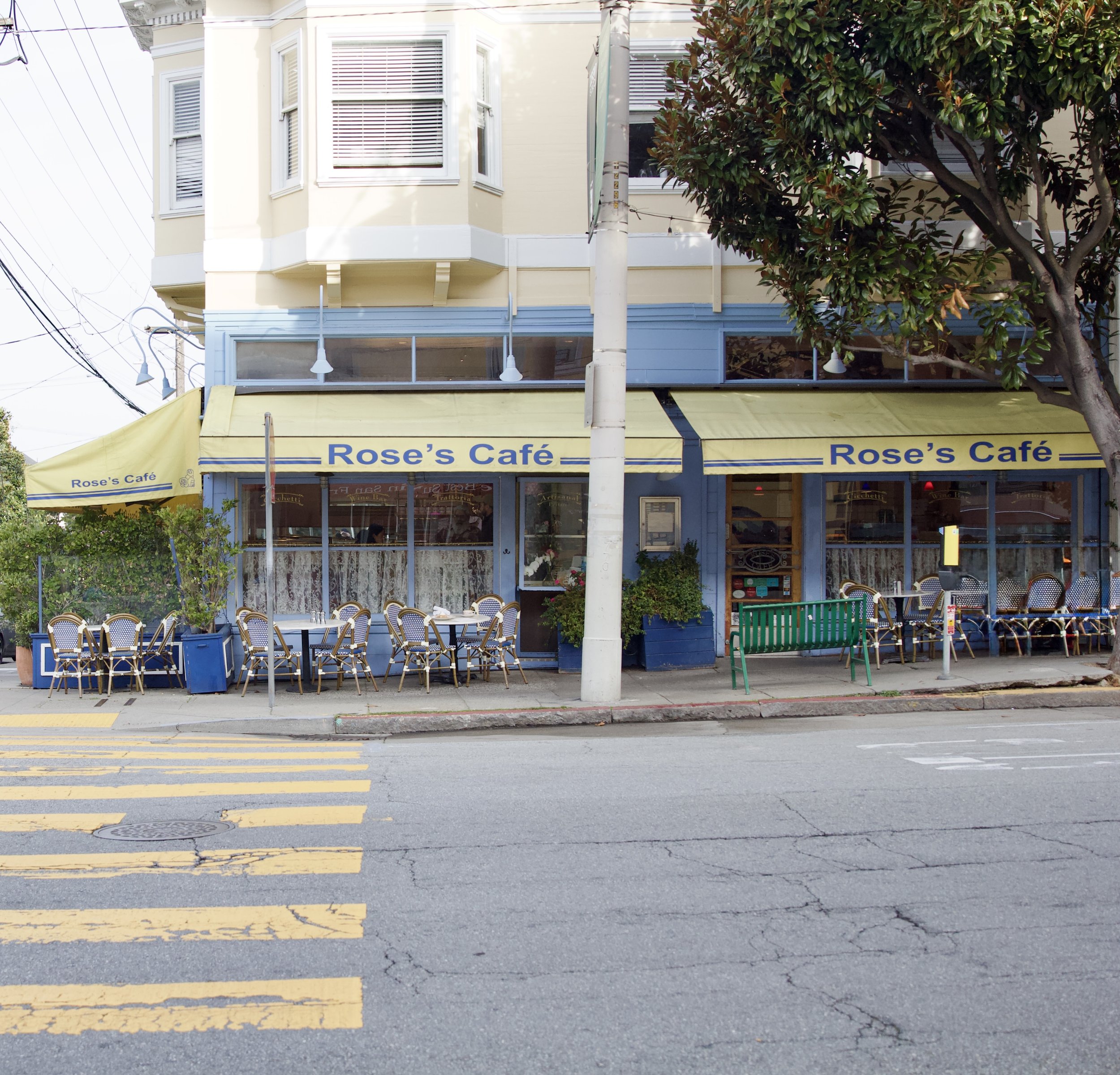 Exterior of Rose's Café with outdoor seating, yellow awnings, and a tree in front on a city street.