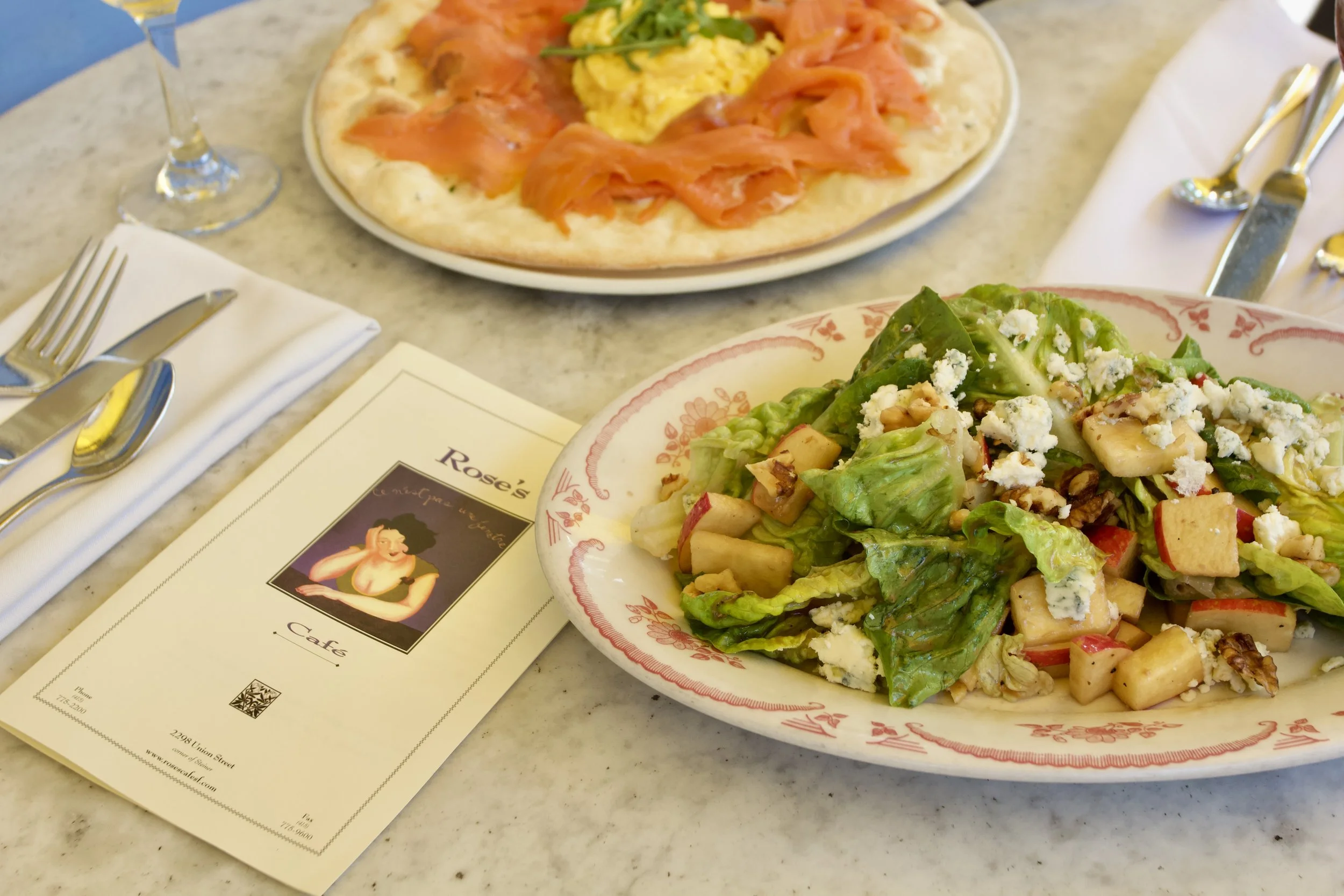 A marble table with a salad with lettuce, apple chunks, blue cheese, and walnuts on a floral plate, a pizza with smoked salmon, scrambled eggs, and green garnish on a white plate, a menu with a woman illustration called Rose's Cafe, and utensils incl