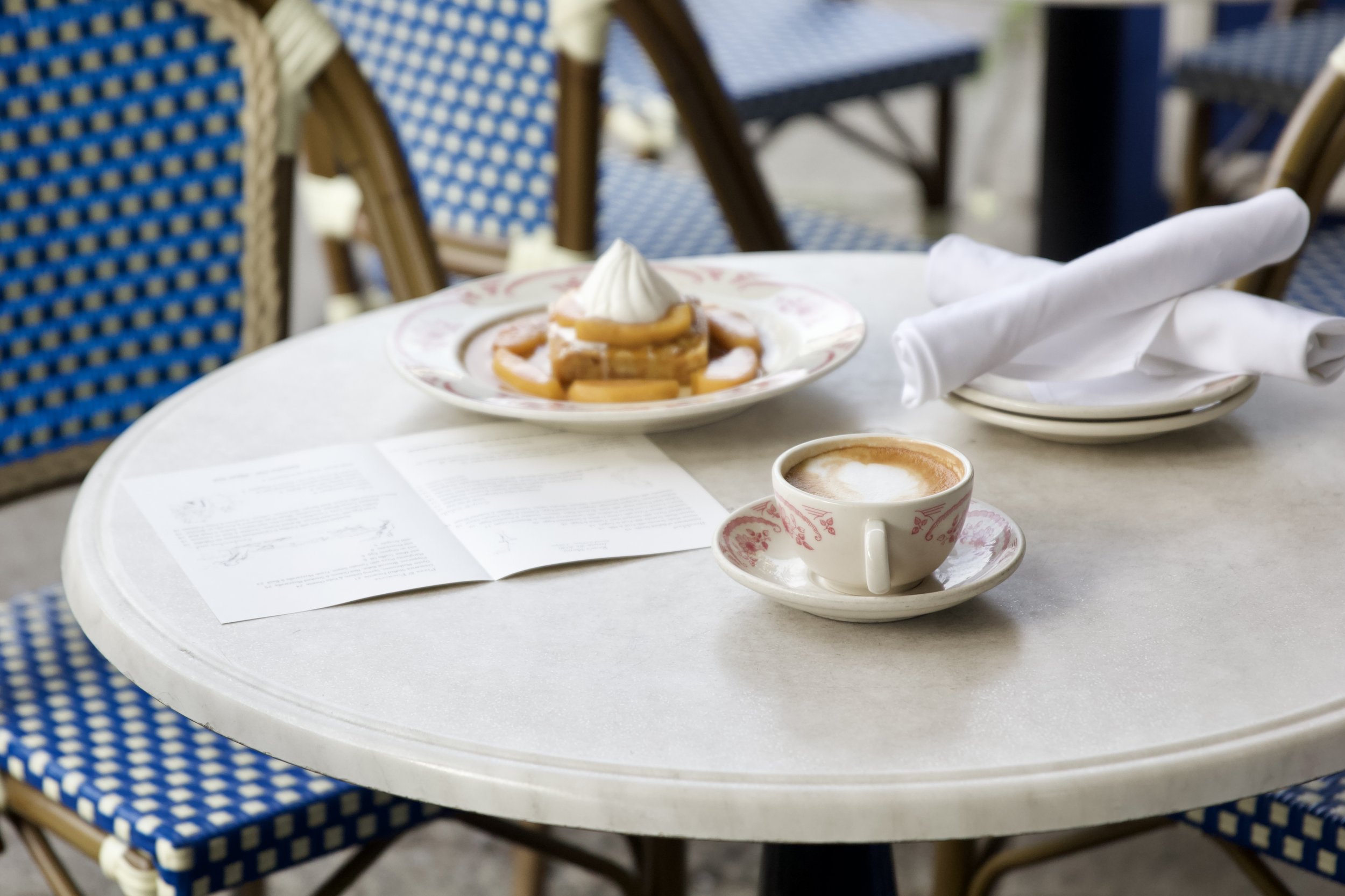 Table with a white tablecloth holding a menu, a cup of coffee on a saucer, a plate with a dessert topped with whipped cream, and a rolled white cloth napkin in an outdoor cafe setting with blue and brown chairs.