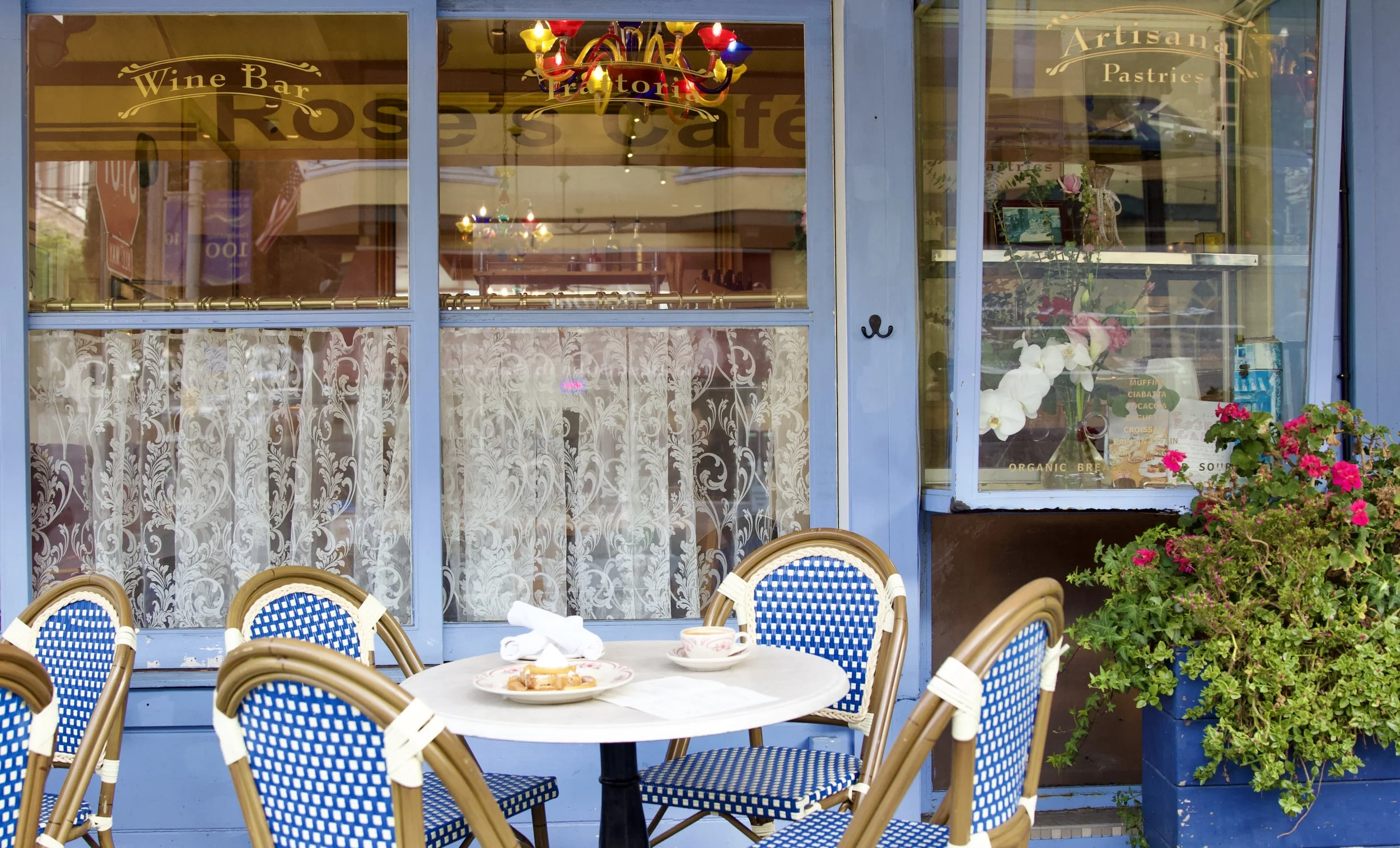 Outdoor café scene with a round white table set for breakfast, surrounded by blue and white wicker chairs. The table has a plate with what appears to be a pastry, a cup, and a napkin. Behind the café, there is a window with lace curtains and a displa