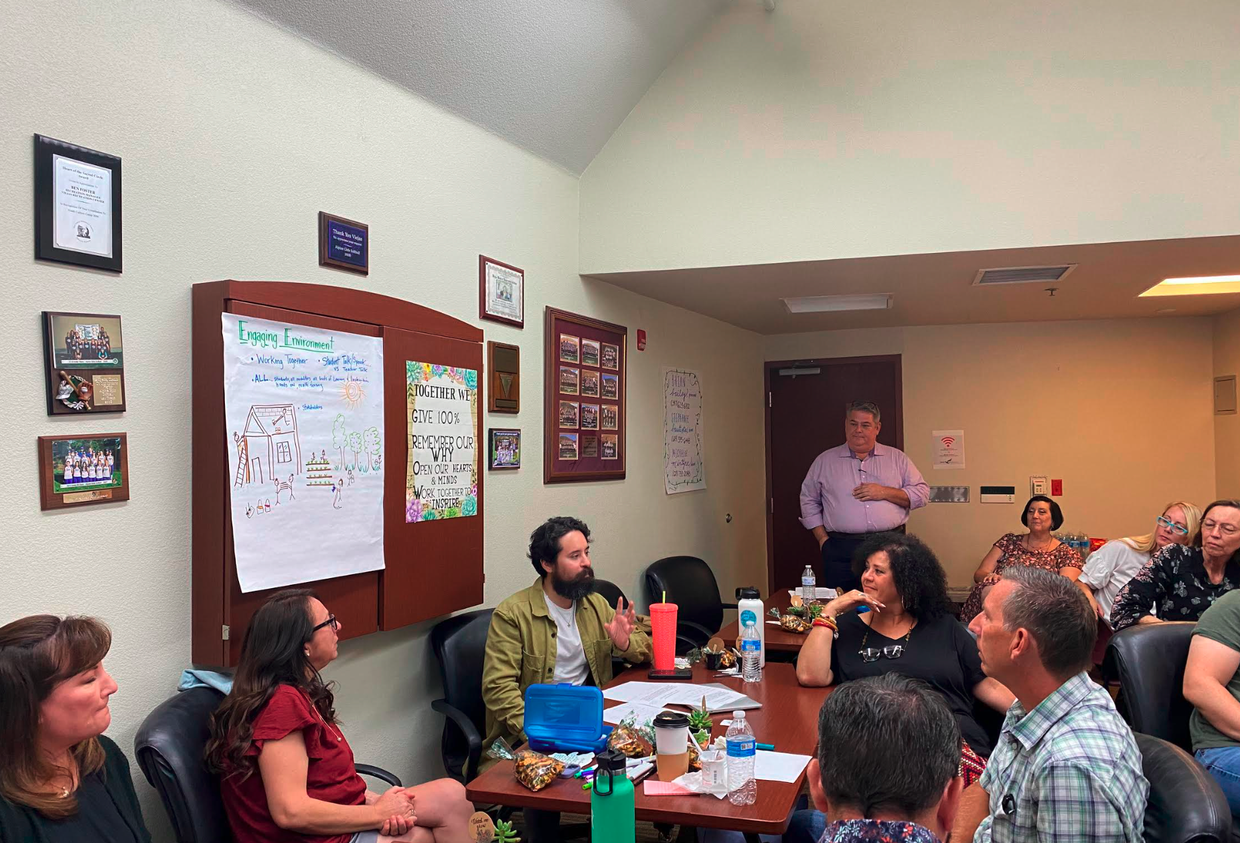 A group of people sitting around a table in a meeting room, with some standing and a man speaking. The walls are decorated with framed certificates and posters, and there is a large whiteboard with drawings and text.