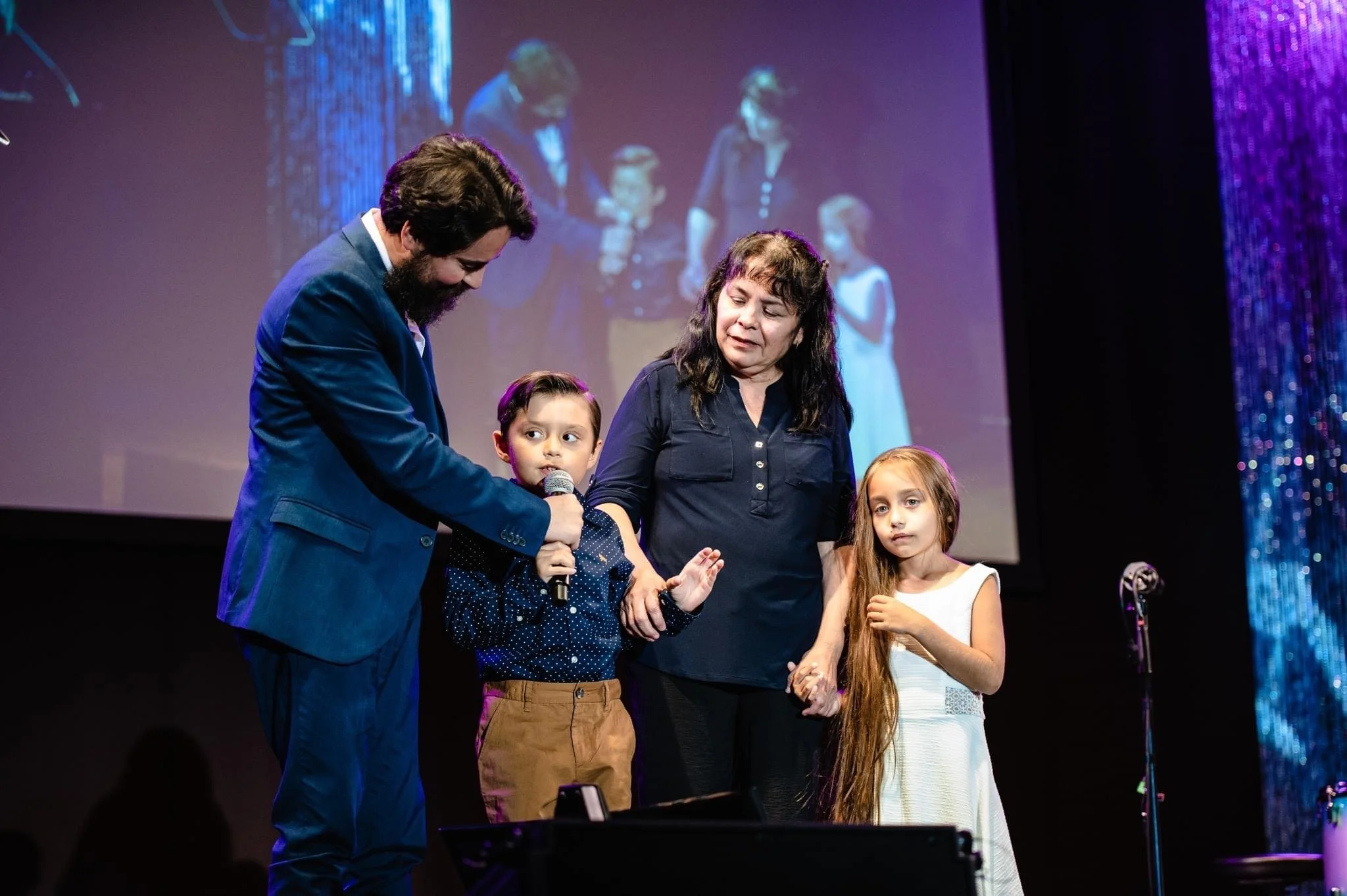 A man in a blue suit holds a microphone for a young boy speaking on stage, with a woman and a girl standing beside them, at an event with a large screen in the background showing a reflection of the scene.