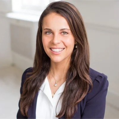 A woman with long brown hair smiling, wearing a navy blazer and white shirt, in an indoor setting.