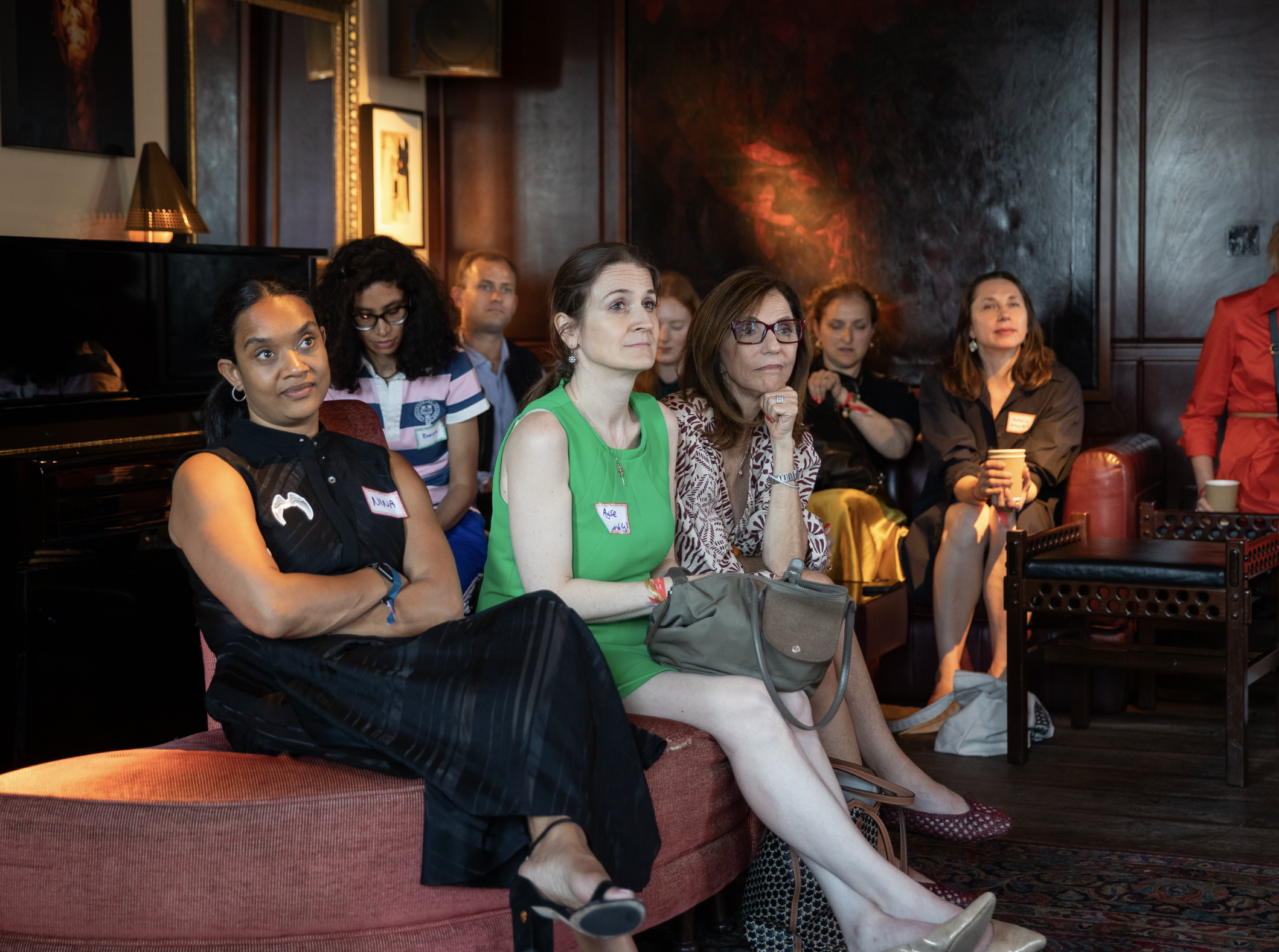 A group of women sitting on red velvet chairs in a dimly lit room, attentively listening, with some holding cups. The room has dark wooden walls and artwork hanging on the wall.