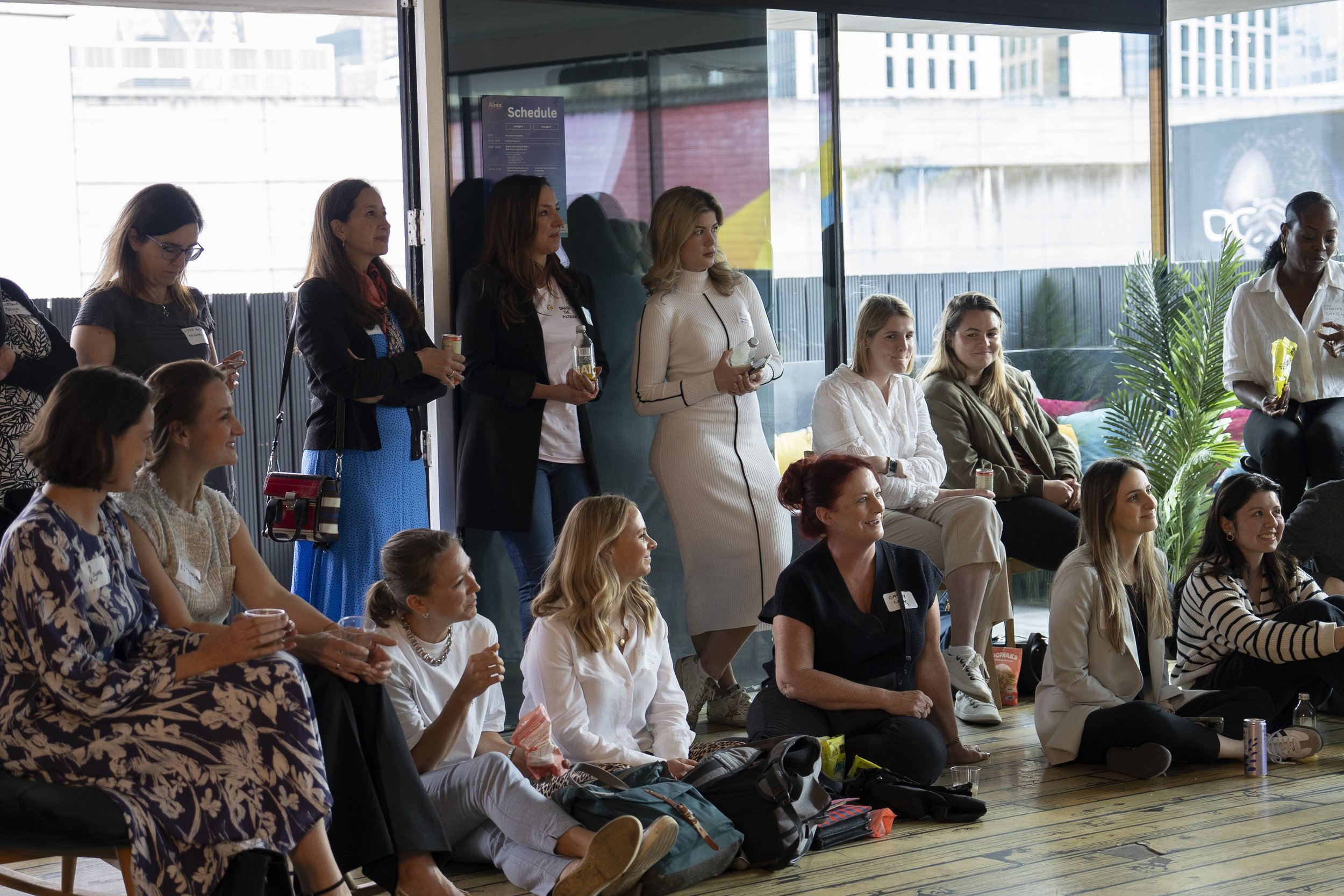 A group of women at a professional or social event, some standing and some seated, in a modern indoor space with large windows and a cityscape view outside.