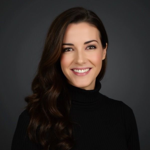 Portrait of a woman with long wavy brown hair, smiling, wearing a black turtleneck against a dark gray background.