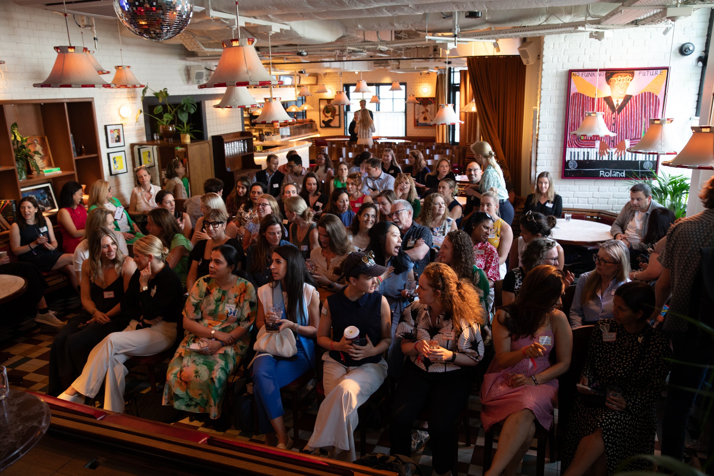 A lively indoor gathering with many people seated and engaging in conversation, set in a warmly lit room with white brick walls, decorative art, and multiple hanging lamps.