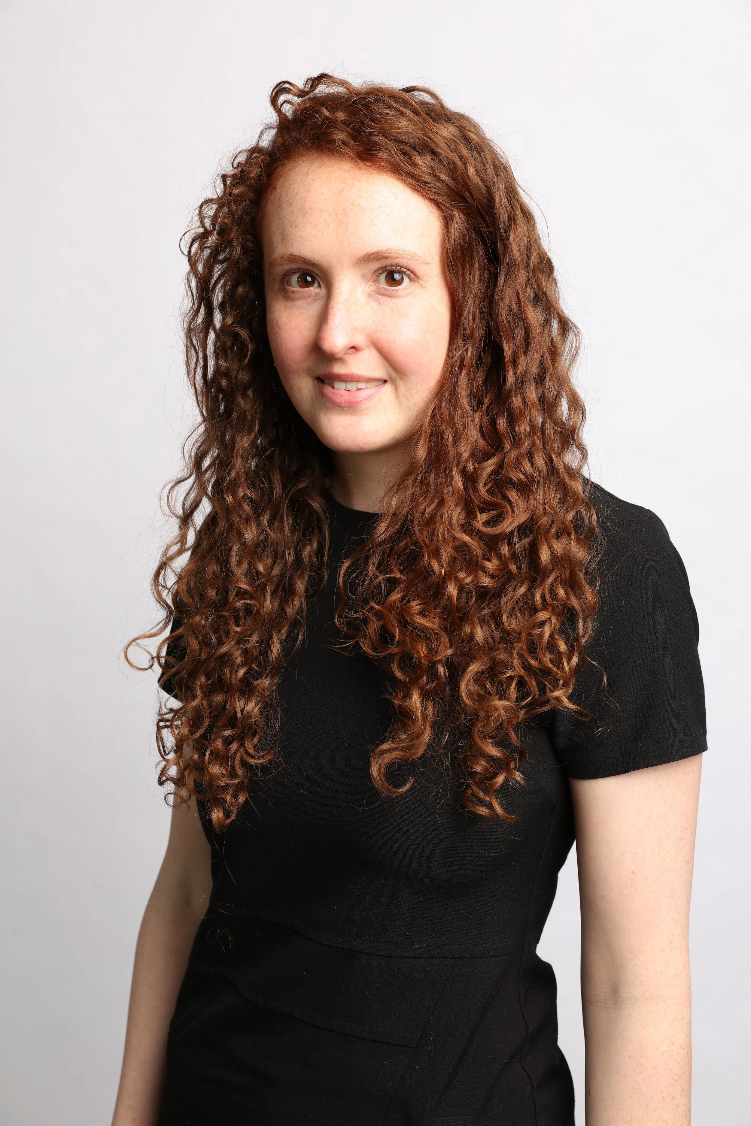 A woman with long, curly auburn hair wearing a black top, smiling and looking at the camera against a plain white background.