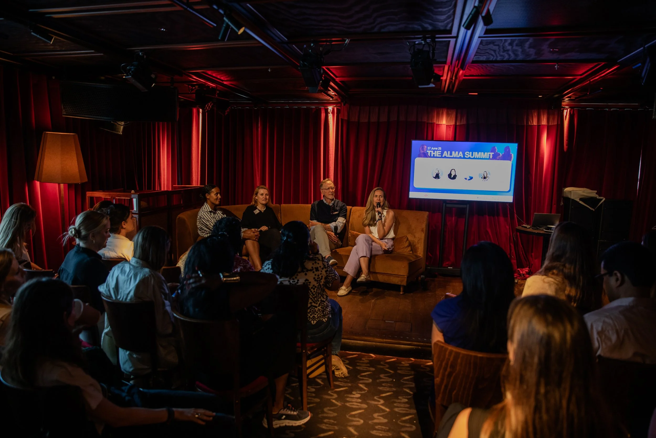 People seated and listening to a panel discussion on stage with a large screen showing event information, in a room with red curtains and dim lighting.
