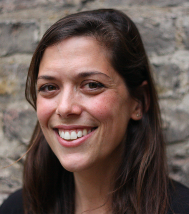Close-up of a woman with long brown hair smiling, standing in front of a stone wall.